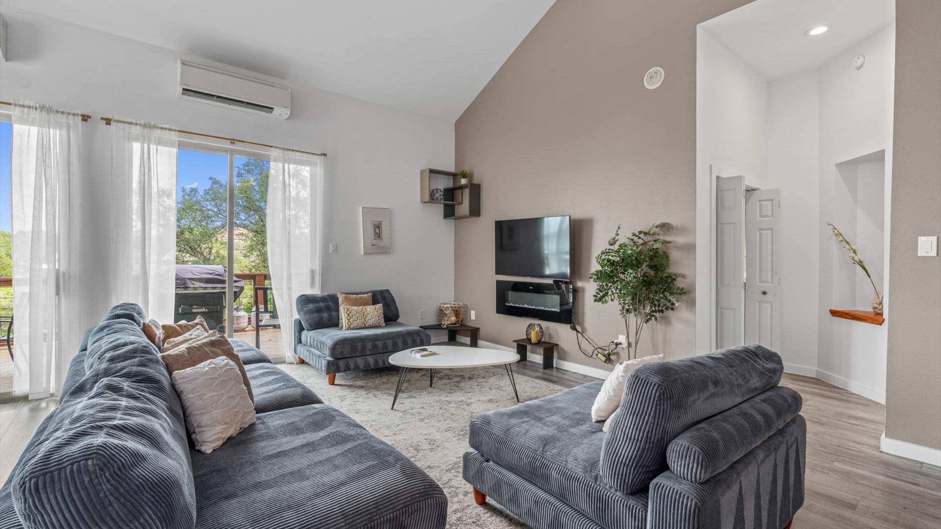 Modern living room featuring a large gray sectional sofa, a wall-mounted TV above an electric fireplace, and a light beige accent wall. Large sliding glass doors lead to an outdoor deck with scenic views.