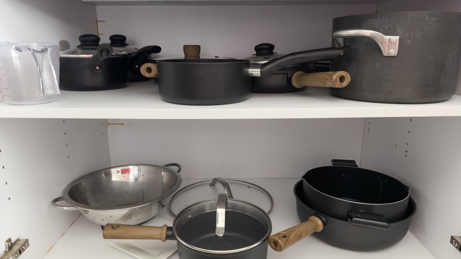 Interior view of a kitchen cabinet revealing a collection of various pots and pans, including saucepans with wooden handles and a colander, neatly stored on white shelves.