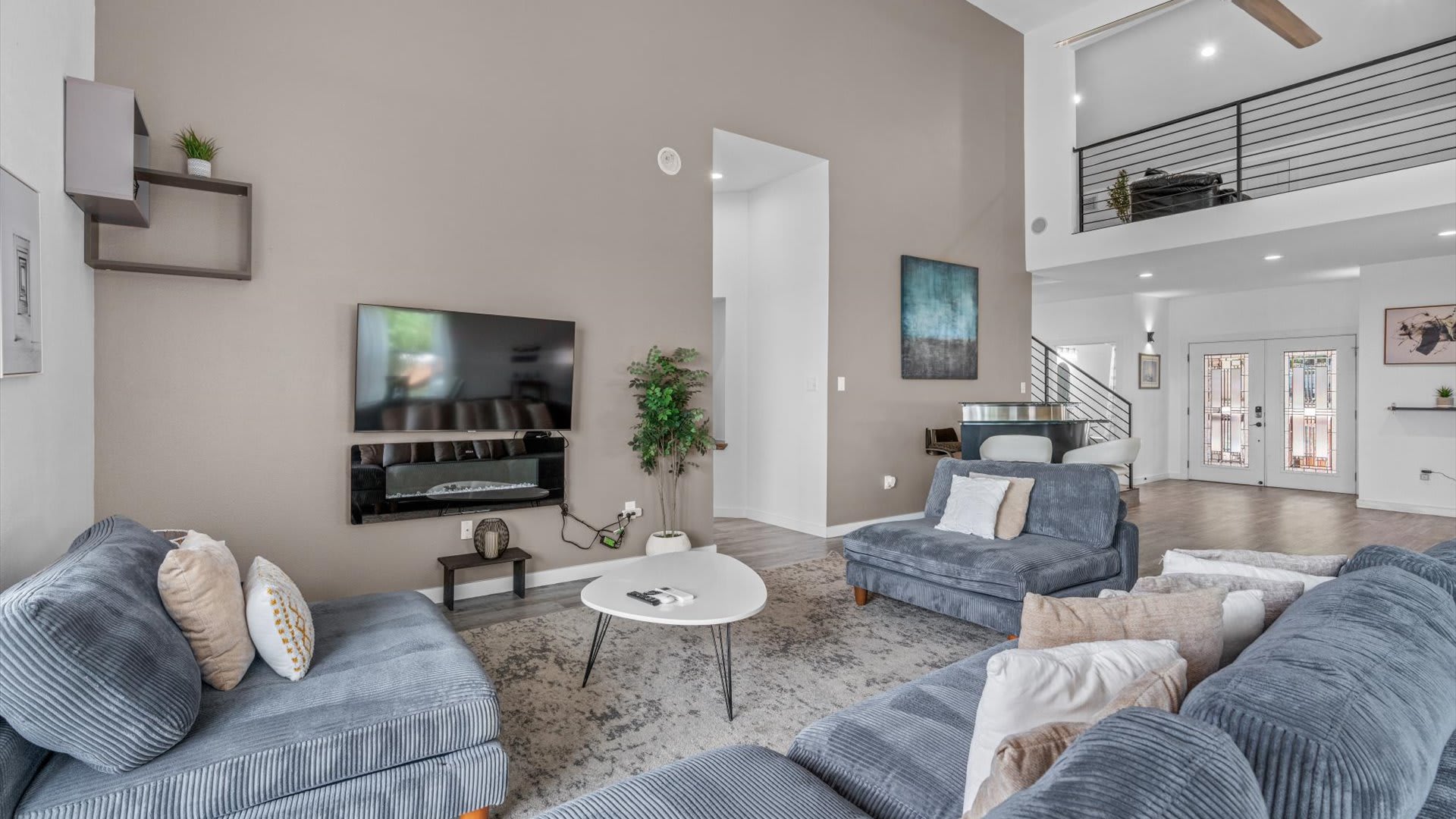 View of the living room from a different angle, showcasing the large gray sectional sofa, a wall-mounted TV, and a light beige accent wall. A bar area with two stools and a staircase leading to an upper level are visible in the background.