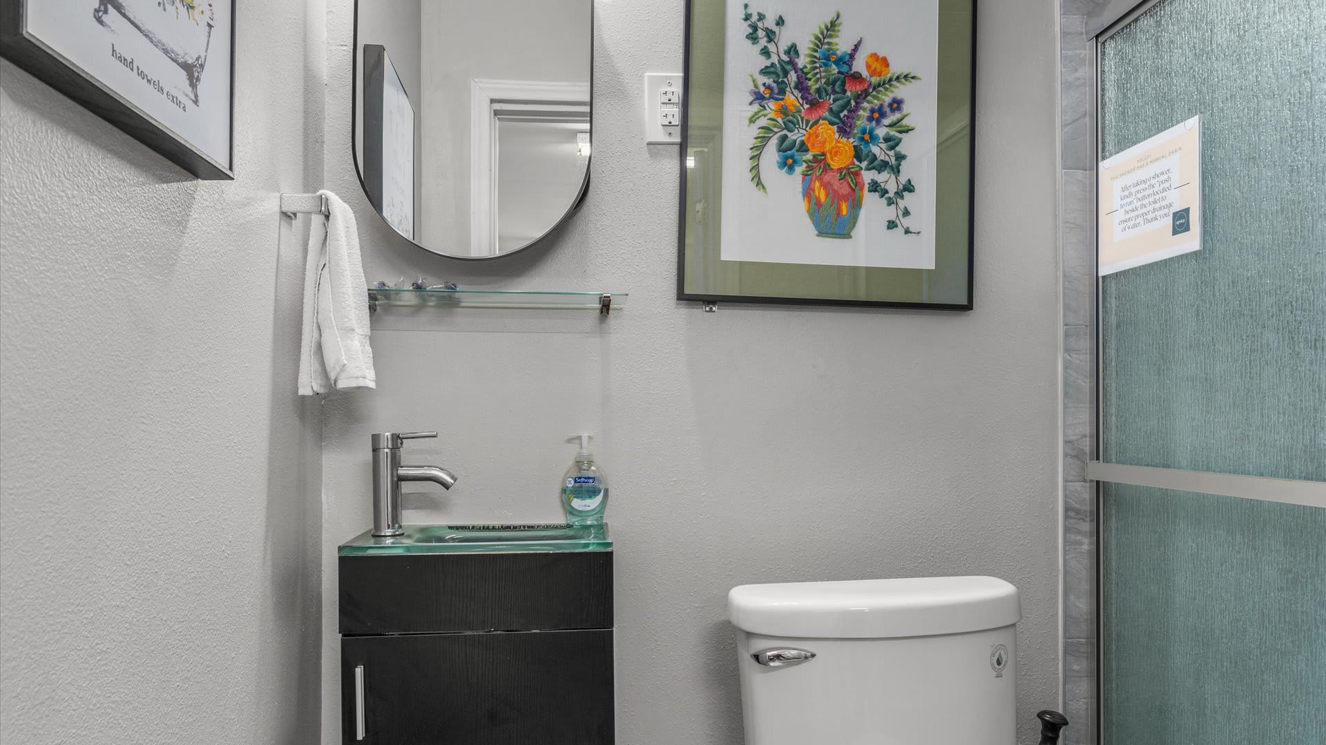 Bright bathroom with a modern black floating vanity and glass counter, a toilet, and a colorful floral artwork on the wall. A round mirror hangs above the vanity.