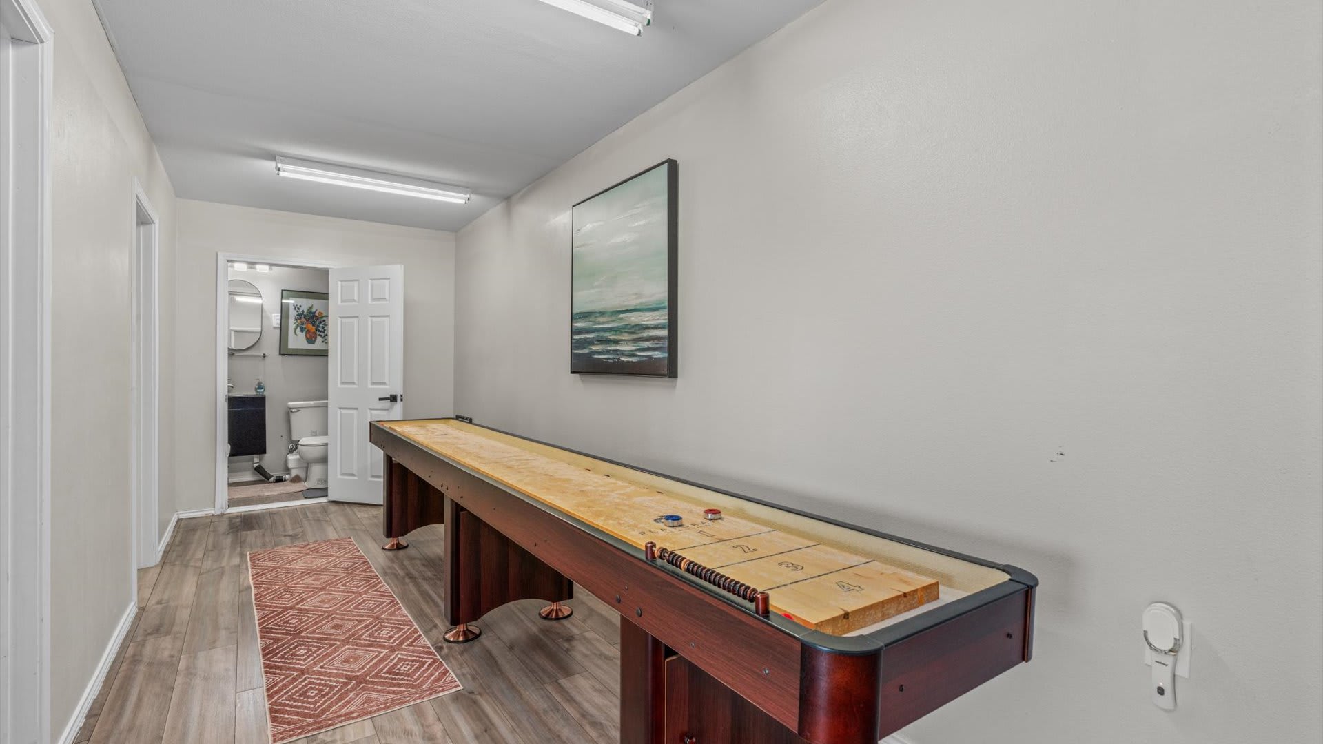A dedicated game room features a long wooden shuffleboard table with pucks, an area rug, and a doorway leading to a bathroom, all set against light-colored walls.
