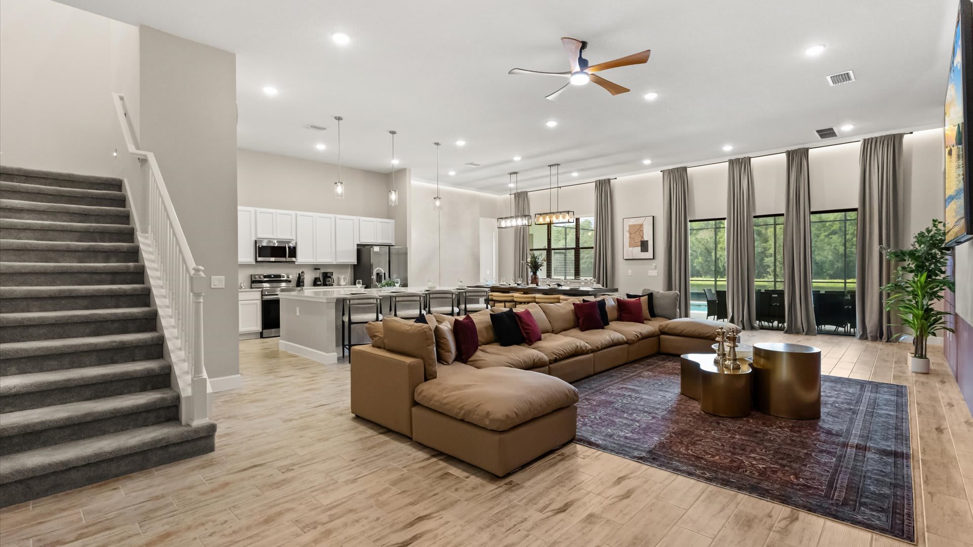 A spacious open-concept living area with a large L-shaped tan sectional sofa, a dark patterned rug, and gold coffee tables. A staircase with gray carpet is visible on the left, leading to upper floors.