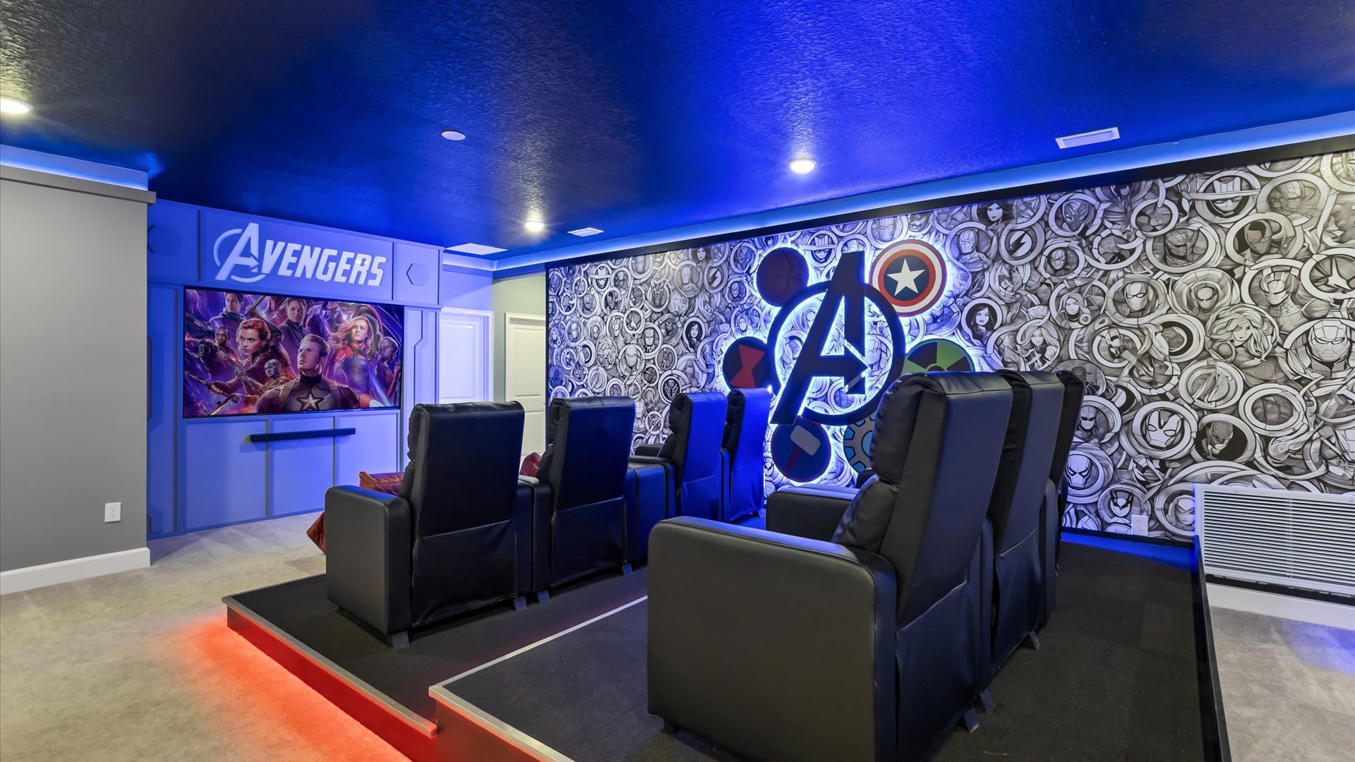 An Avengers-themed media room features comfortable black reclining chairs arranged on tiered platforms facing a large screen displaying a movie. The walls are decorated with comic book art and a large illuminated Avengers logo, all under blue accent lighting.