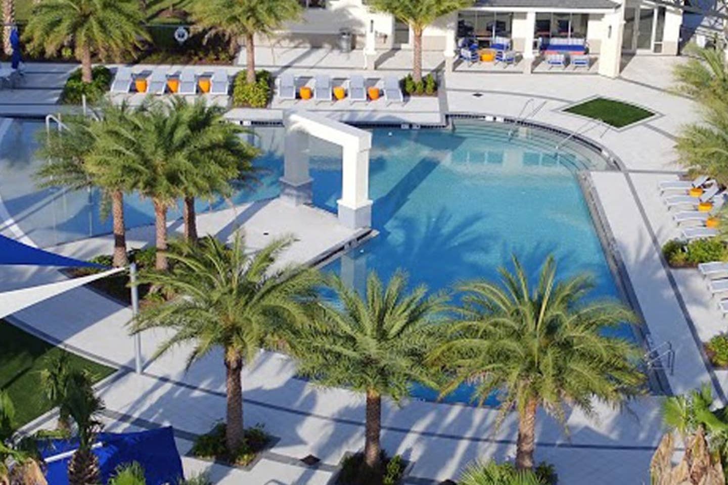 Large communal swimming pool surrounded by palm trees and lounge chairs, with a white archway feature in the center. The pool area is paved with light-colored tiles.