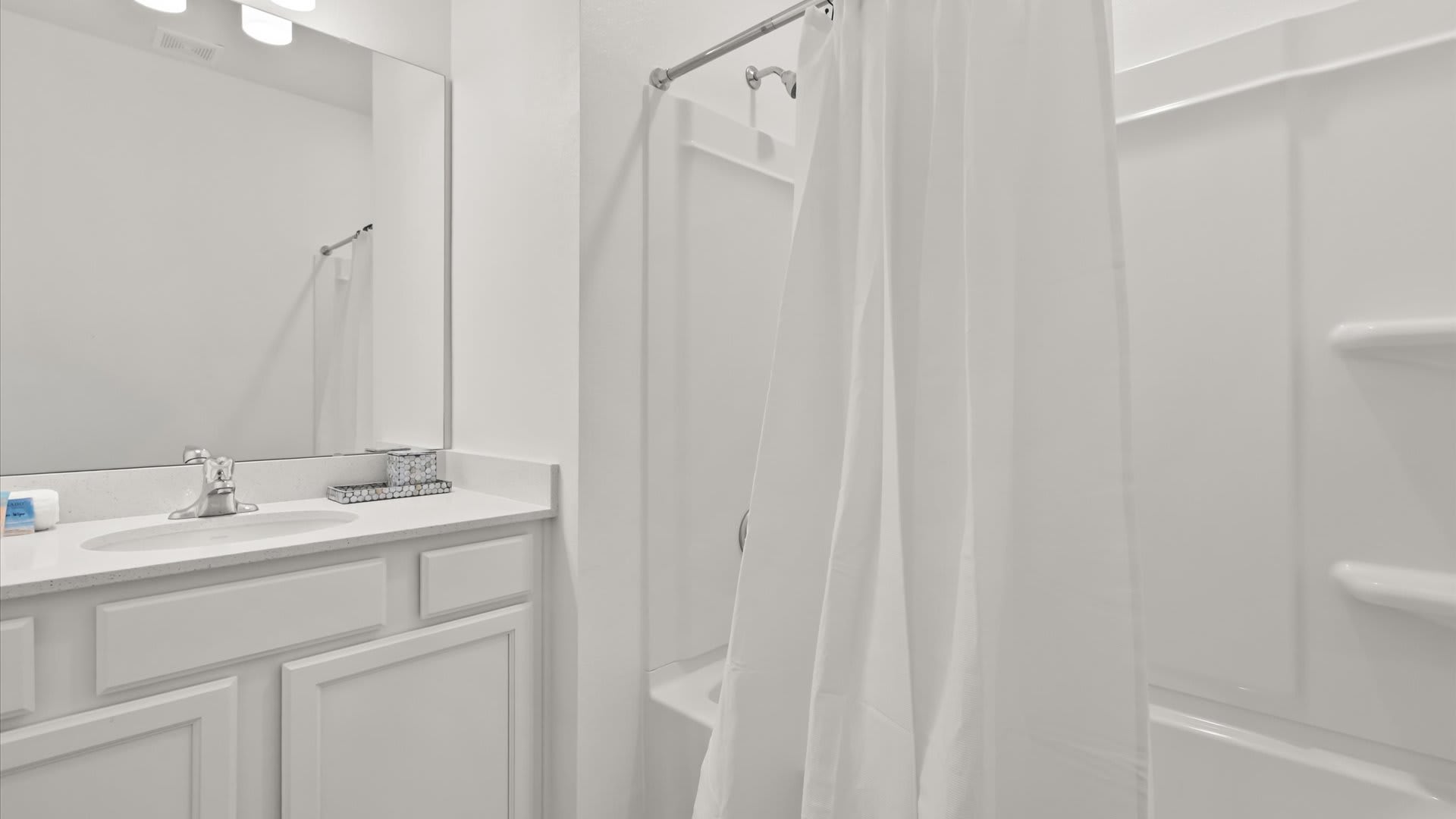 Bright white bathroom featuring a single vanity with a white countertop, chrome faucet, and a large mirror. A shower/tub combo with a white shower curtain is visible on the right.
