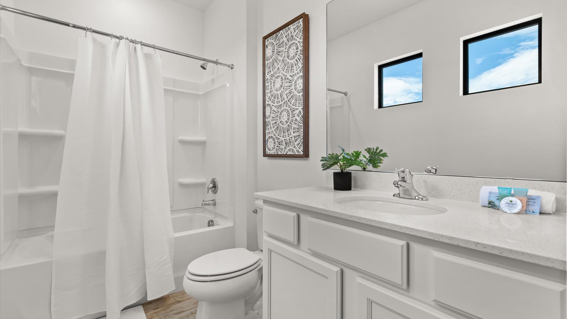 Clean white bathroom featuring a single vanity with a white countertop, chrome faucet, and a shower/tub combination with a white curtain. Two small square windows are above the toilet.