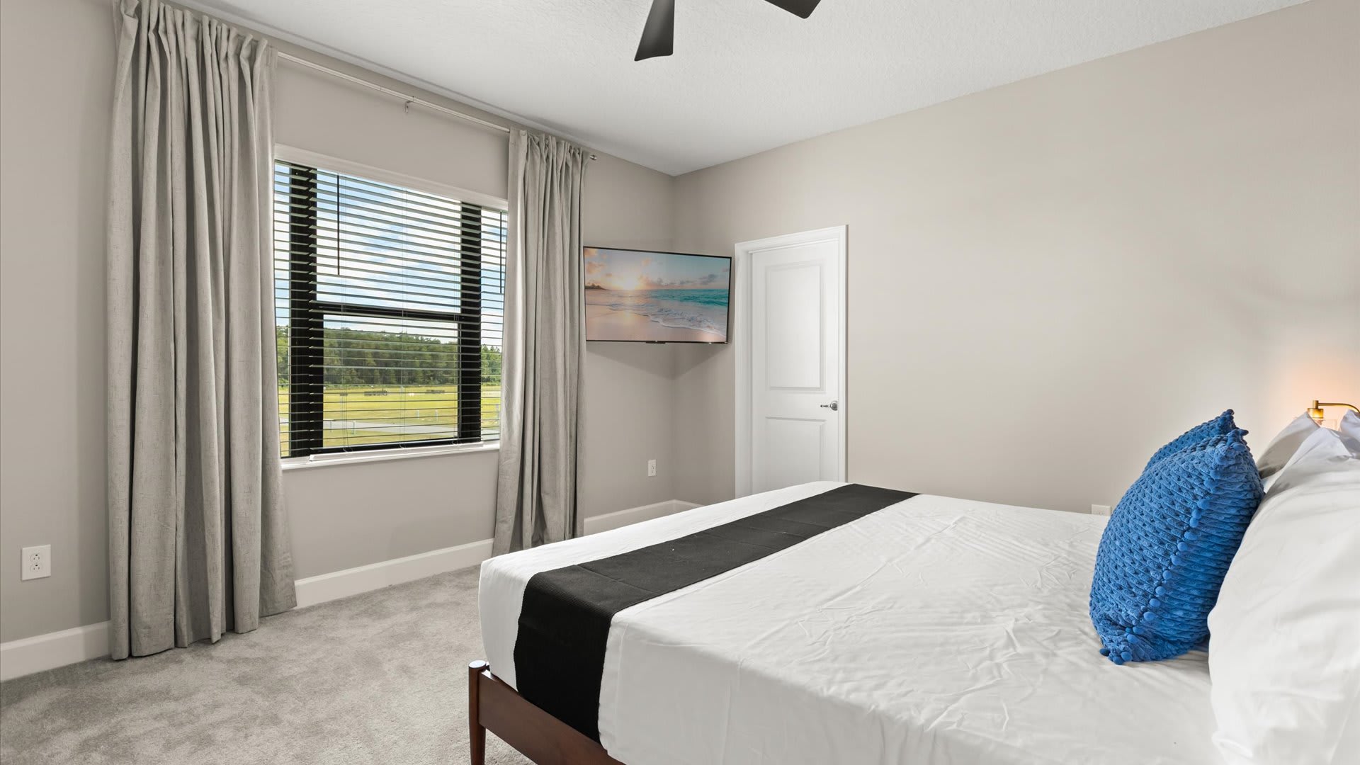 Bright bedroom with a large window offering a view of greenery, covered by blinds and light gray curtains. A wall-mounted flat-screen TV is positioned opposite the bed, above a dark dresser.