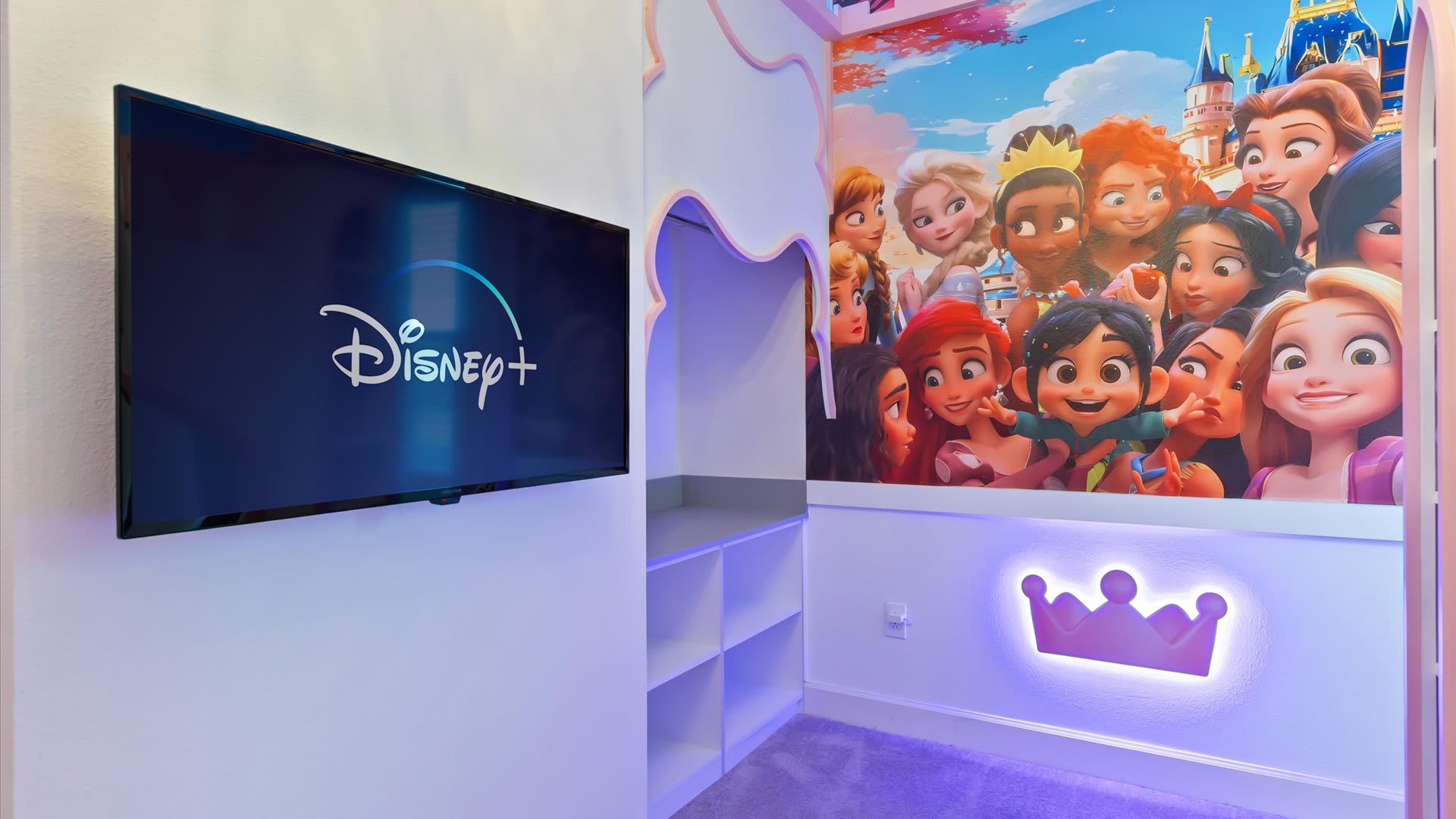 Another perspective of the Princess themed bedroom, showcasing a wall-mounted TV displaying the Disney+ logo, positioned next to the vibrant Disney Princess mural and the illuminated crown decor.