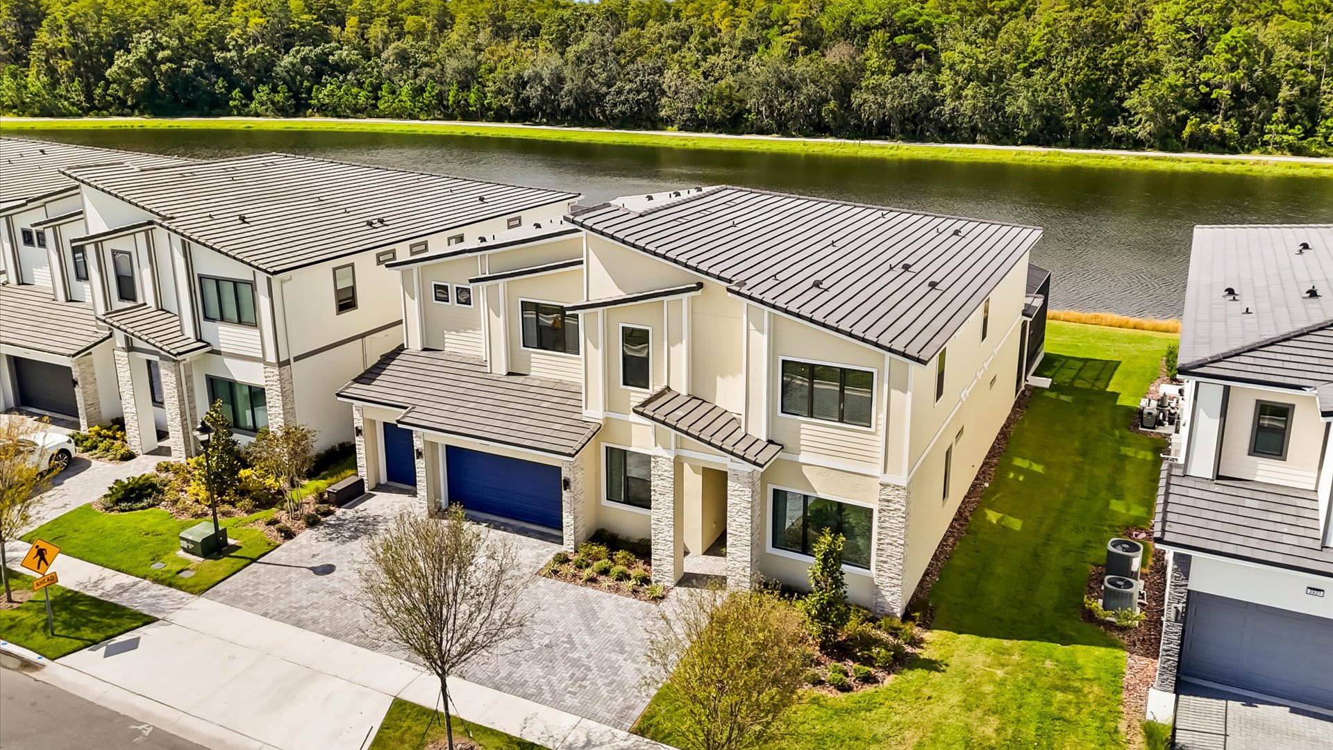 Aerial view of a modern two-story vacation home with a light-colored exterior and dark roof, situated next to a serene lake. The property features a paved driveway and manicured lawn.
