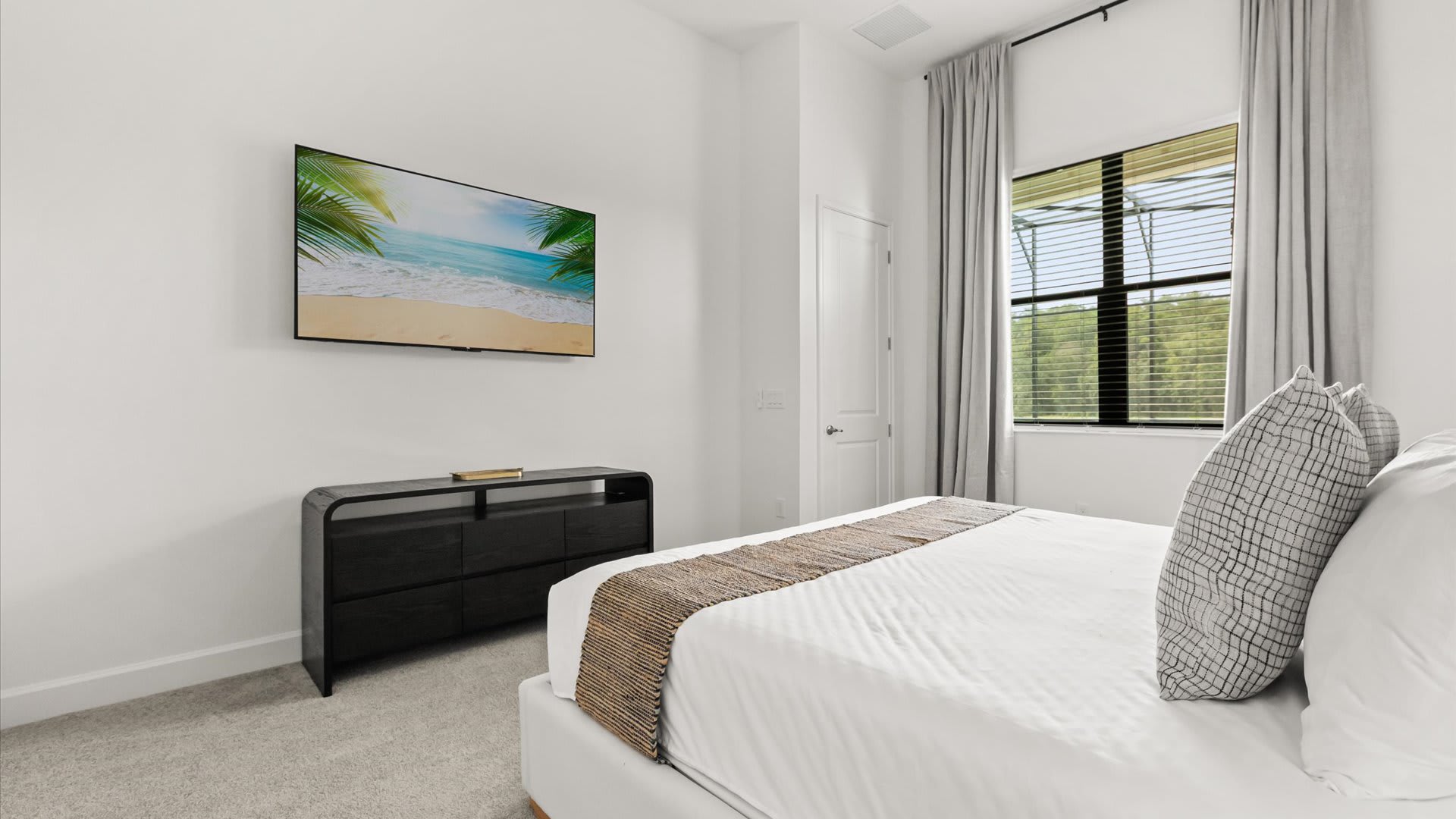 Contemporary bedroom with a large window providing a view of trees, dressed with light gray curtains. A wall-mounted flat-screen TV is positioned above a dark, low-profile dresser.
