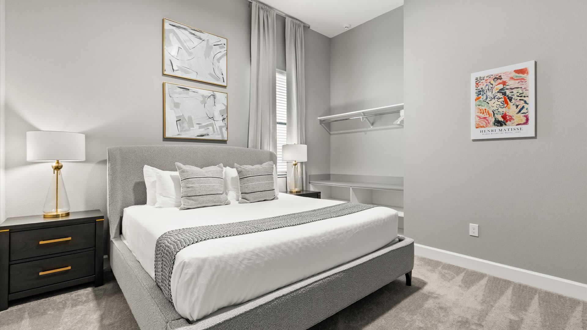 A stylish bedroom with light gray walls, a gray upholstered bed with white and gray bedding, and two black nightstands with gold-accented lamps. Two abstract artworks hang above the bed, and an open closet with shelving is visible.