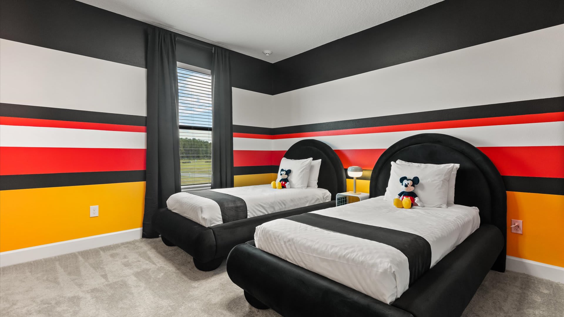 Vibrantly themed bedroom with black, white, red, and yellow striped walls, featuring two black upholstered twin beds with white bedding and Mickey Mouse plush toys. A window with horizontal blinds offers an outdoor view.