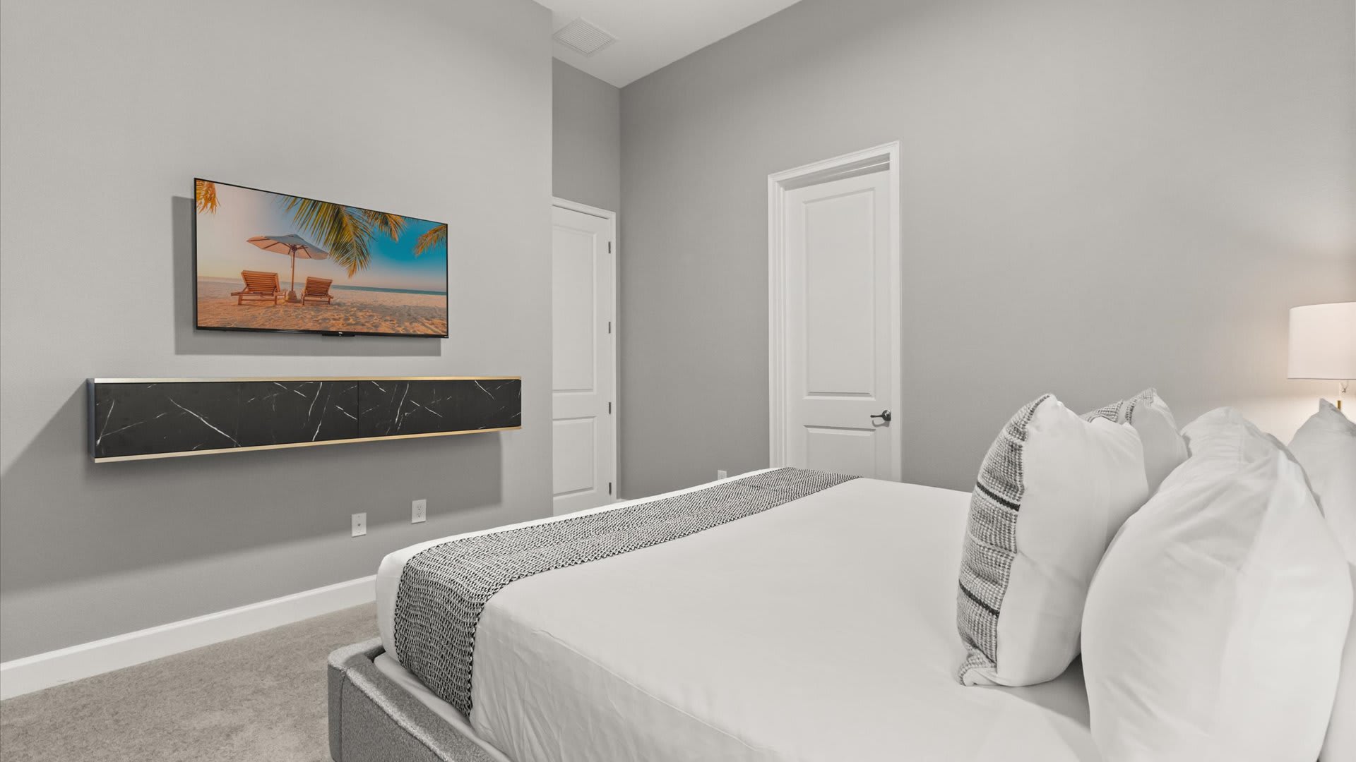 A modern bedroom with light gray walls, a gray upholstered bed with white and gray bedding, and a wall-mounted TV displaying a beach scene. A black and gold floating media console is positioned beneath the TV.