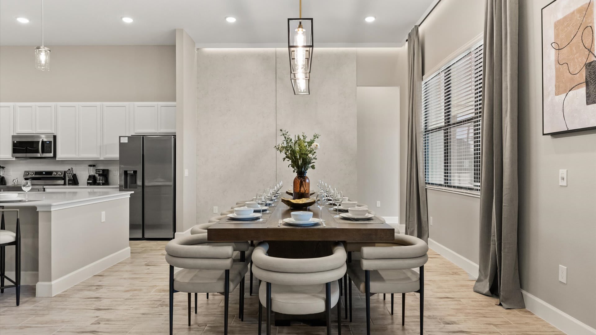 Elegant dining room with a long dark wood table set for multiple guests, surrounded by modern upholstered chairs. A linear chandelier hangs above, and large windows with blinds and drapes offer natural light.
