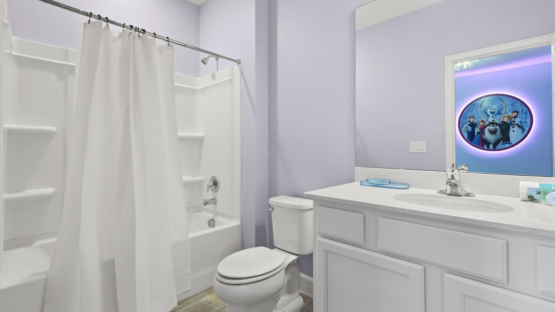 A light purple bathroom features a white vanity with a single sink and a large mirror reflecting the 'Frozen' themed room. It includes a white toilet and a shower/tub combo with a white curtain.