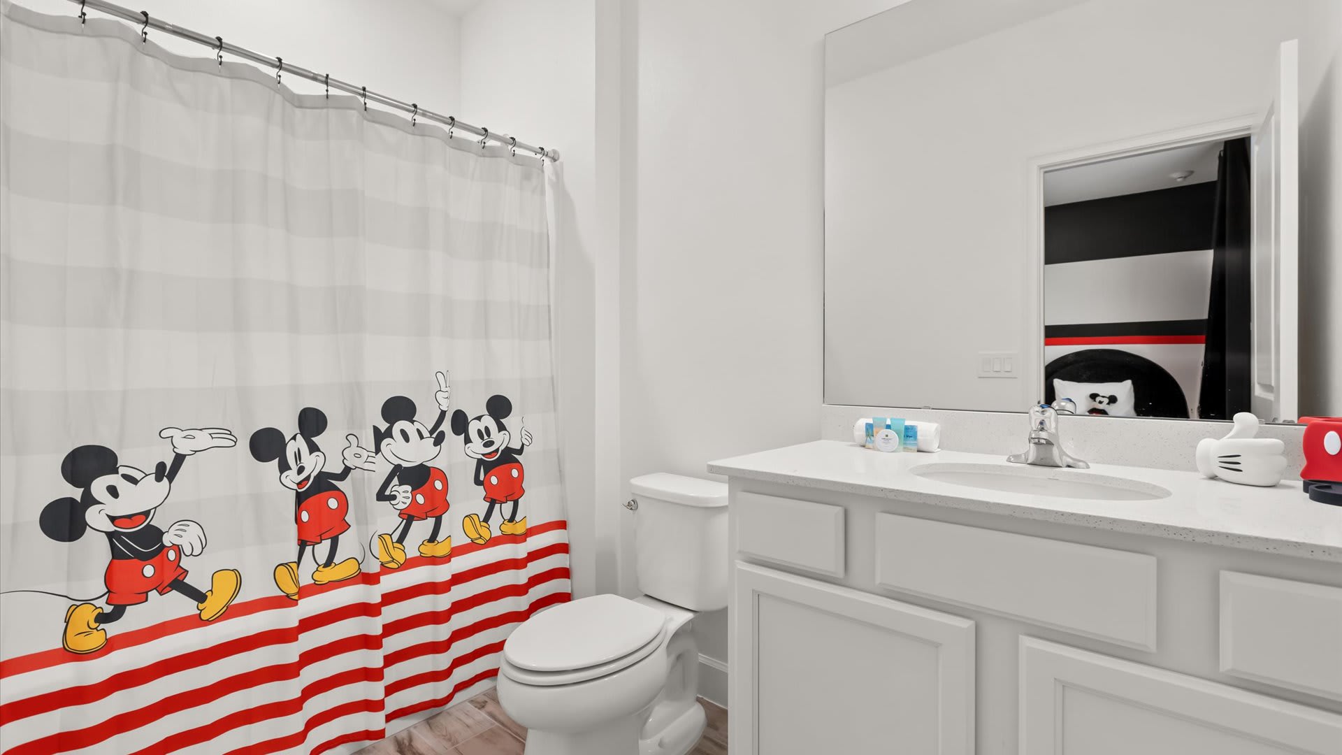 White bathroom with a single vanity, white countertop, and a shower/tub combo featuring a playful red and white striped Mickey Mouse shower curtain. The mirror reflects the Mickey Mouse themed bedroom.