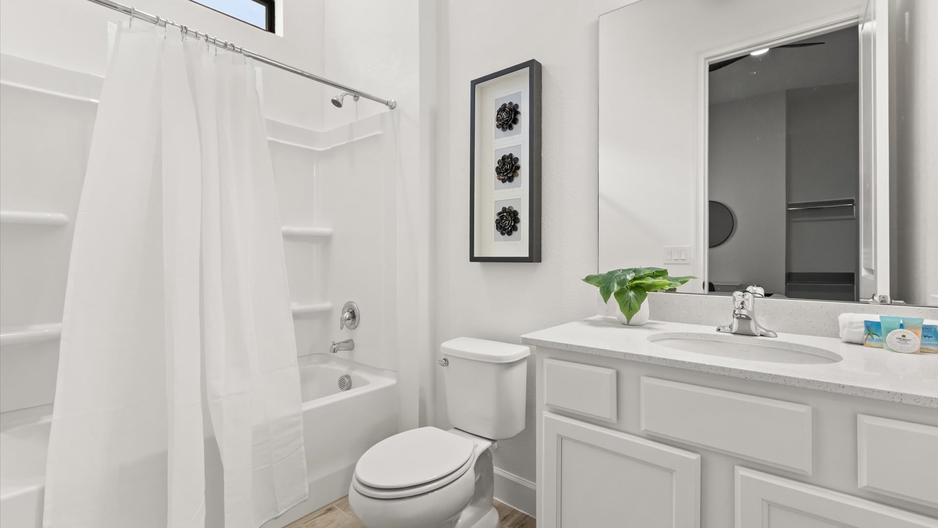 A bright white bathroom featuring a shower-tub combo with a white curtain, a toilet, and a vanity with a white countertop, sink, and mirror. Three framed black floral artworks adorn the wall above the toilet.