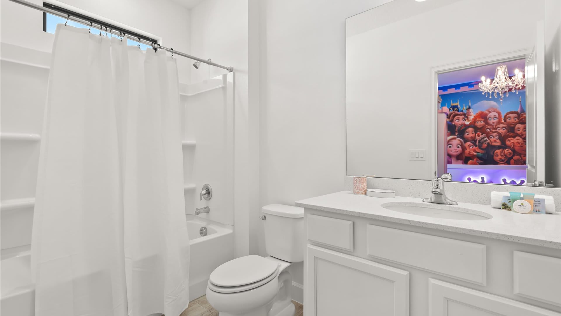 White bathroom with a single vanity, white countertop, and a shower/tub combo with a white curtain. The mirror reflects the Princess themed bedroom with its colorful mural and chandelier.