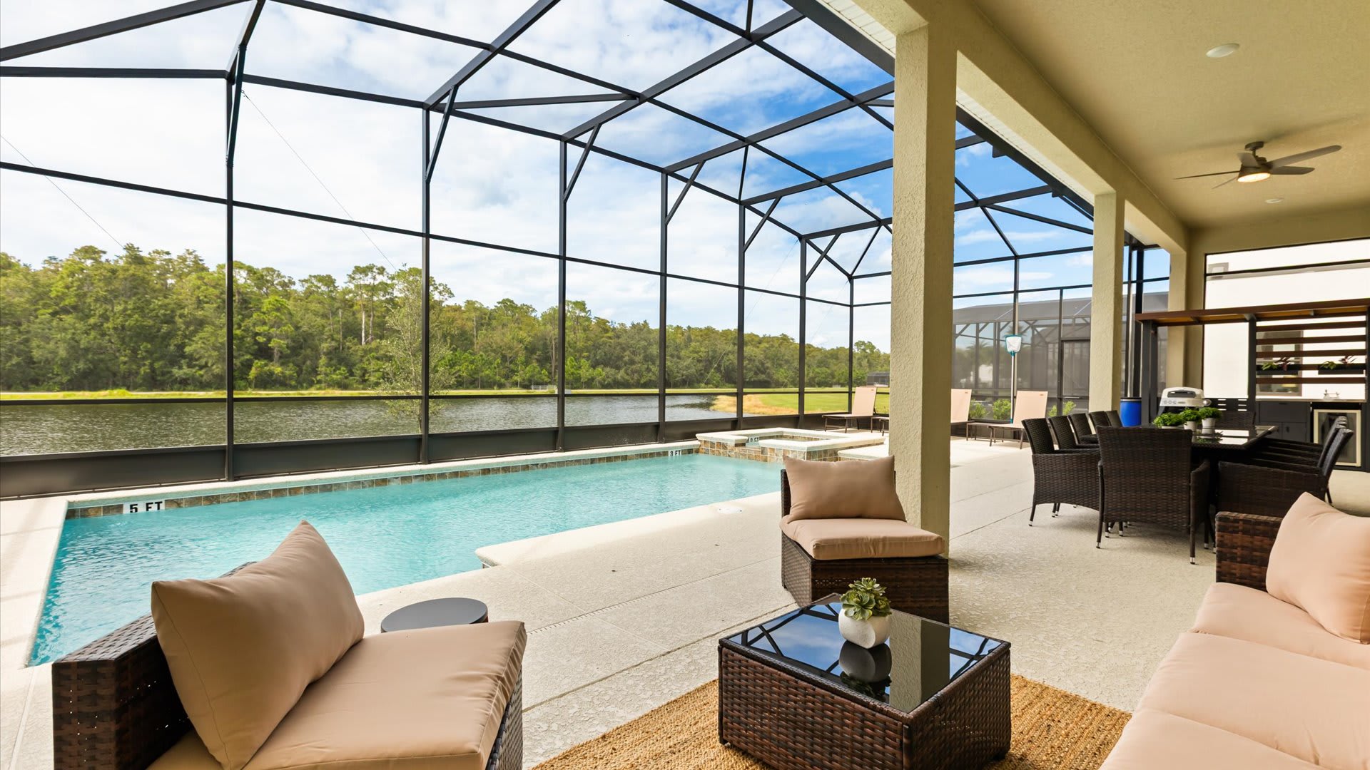 A comfortable outdoor seating area on a covered patio overlooks a screened-in private pool and a serene lake surrounded by lush trees. The patio features wicker chairs and a coffee table, providing a relaxing space with a scenic view.