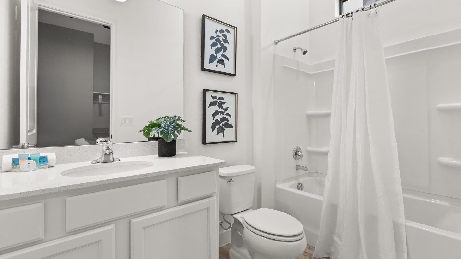 A clean white bathroom with a shower-tub combo, a toilet, and a vanity with a white countertop and sink. Two framed botanical prints in blue and black hang above the toilet, adding a touch of color.