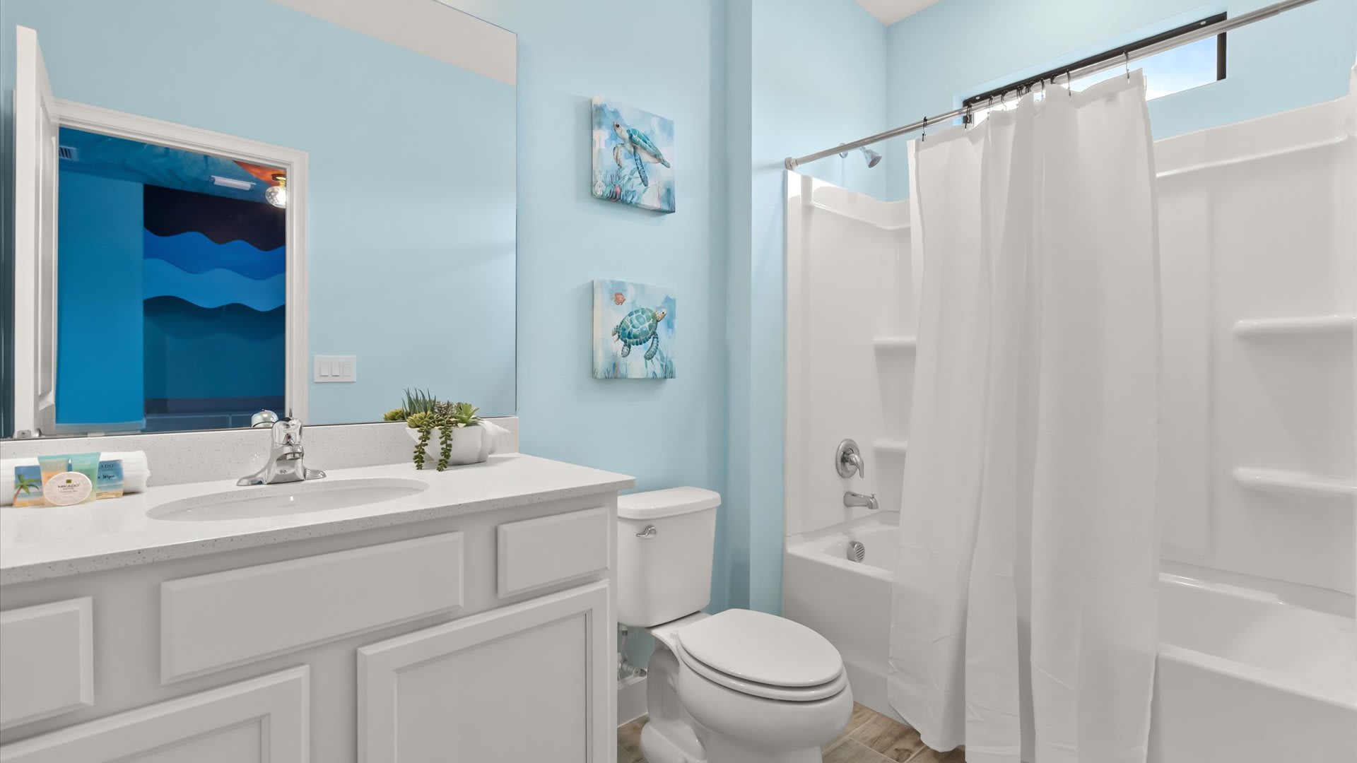 A light blue bathroom features a white vanity with a single sink and a large mirror reflecting the themed bedroom. It includes a white toilet and a shower/tub combo with a white curtain.