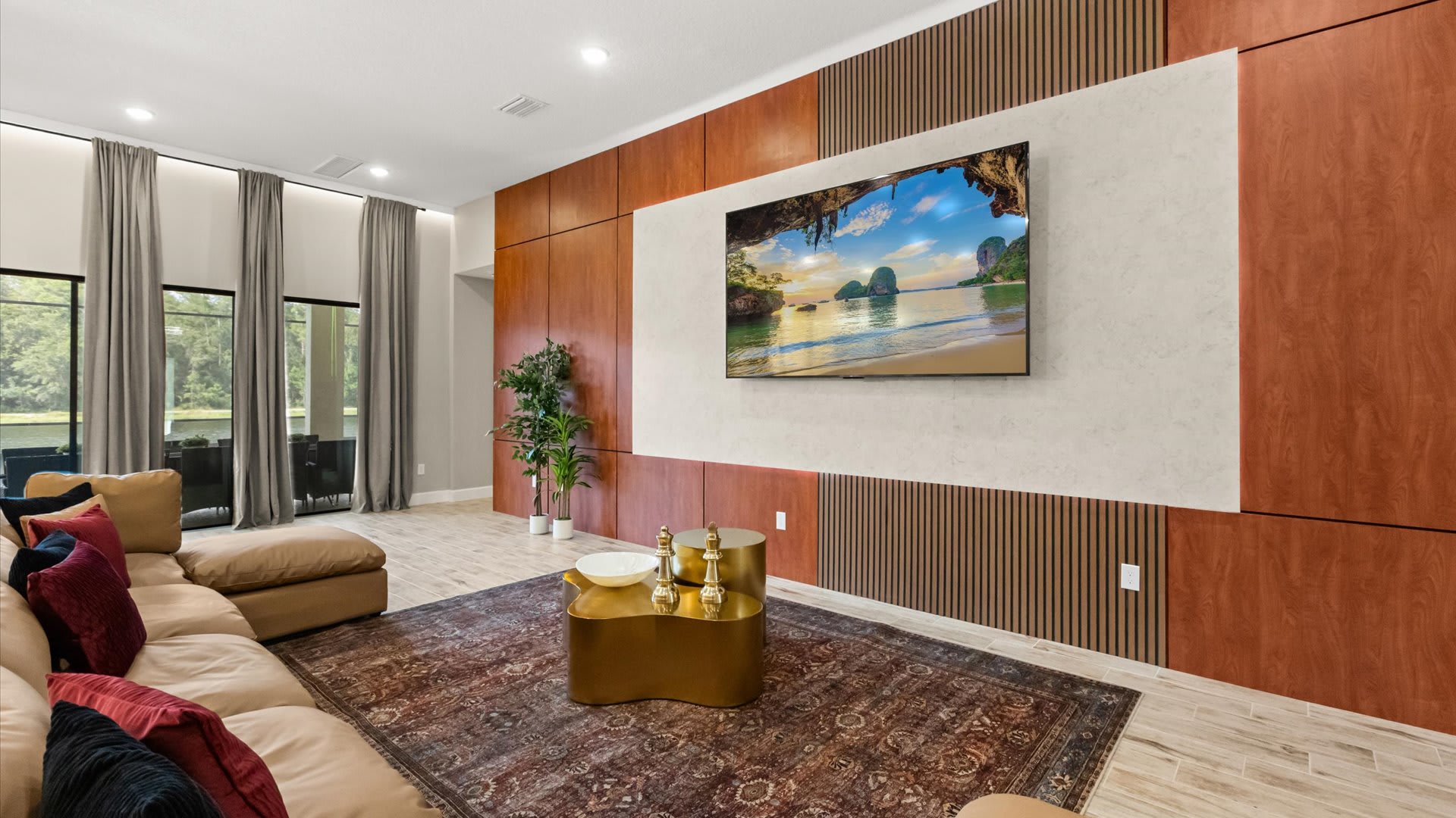 Modern living room featuring a large wall-mounted TV against a textured accent wall with wood paneling. A tan sectional sofa with colorful throw pillows and gold coffee tables sit on a dark patterned rug.