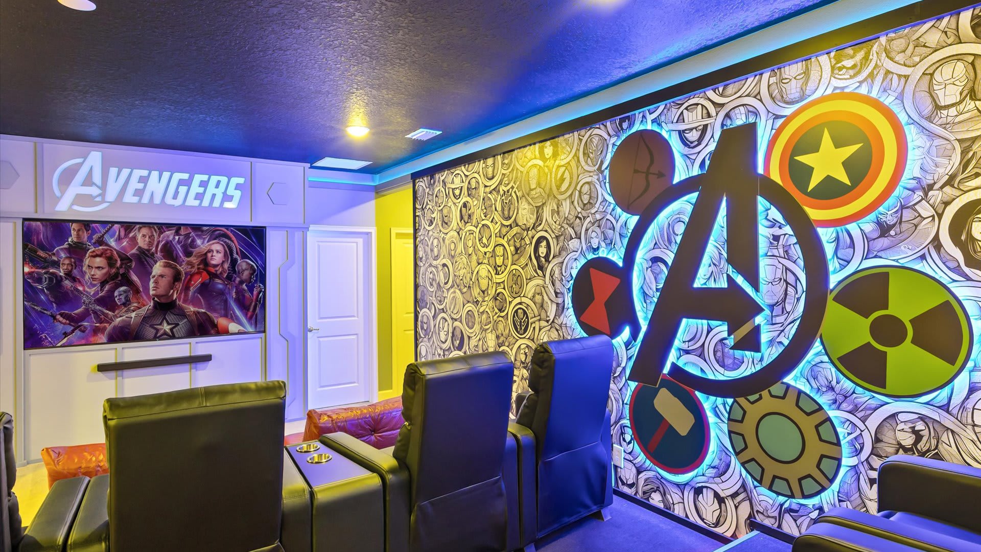 A close-up view of the Avengers-themed media room highlights the large illuminated logo and comic book wallpaper on the wall. Comfortable reclining chairs are visible, set on tiered platforms, ready for a cinematic experience.