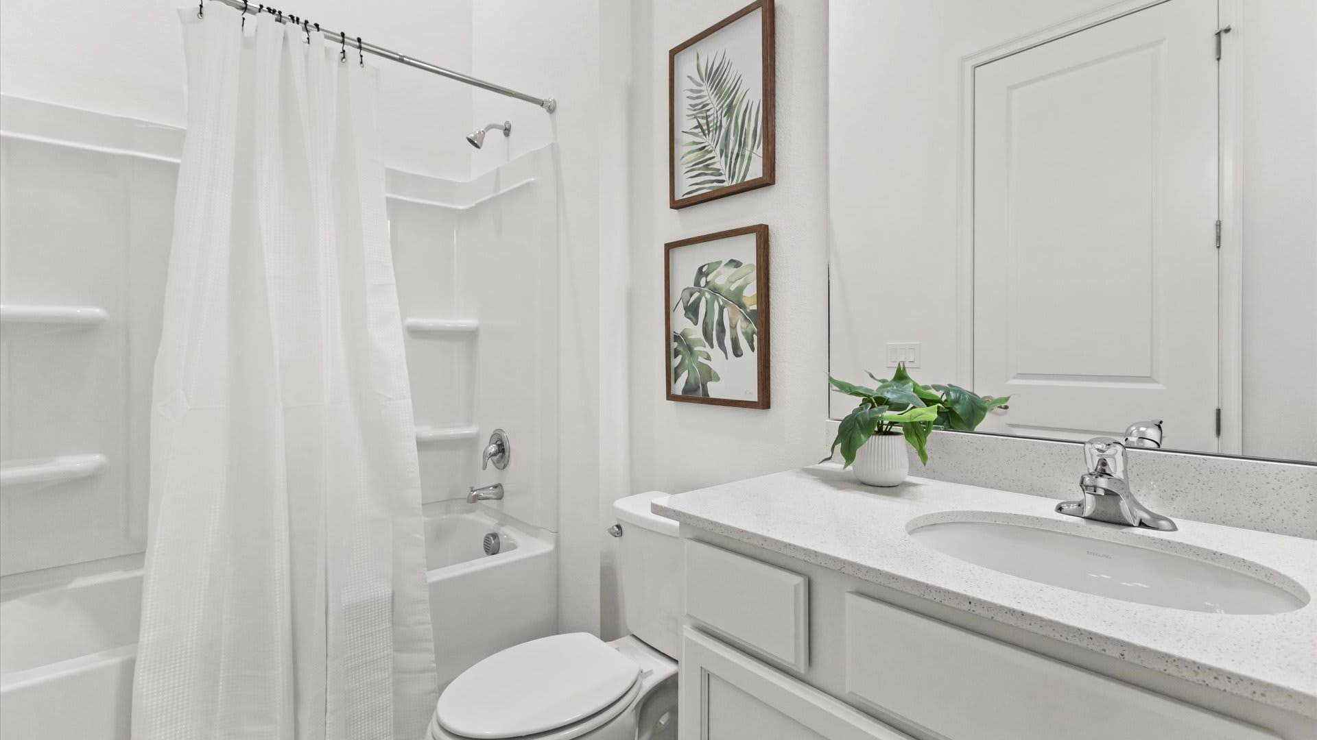 A clean white bathroom featuring a shower-tub combo with a white curtain, a toilet, and a vanity with a white countertop and sink. Two framed green botanical prints hang above the toilet, adding a natural touch.