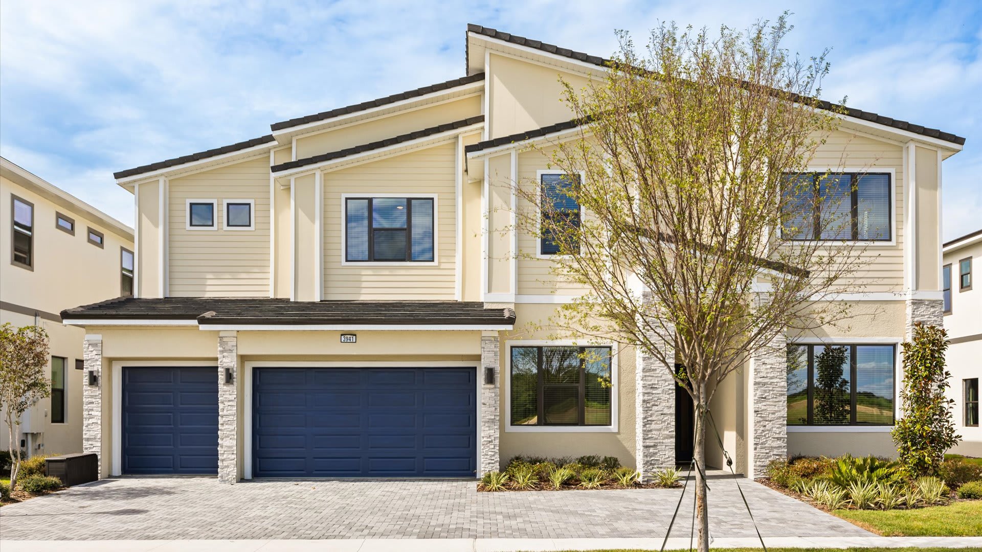 Modern two-story vacation home with light yellow siding and stone accents, featuring two dark blue garage doors and a paved driveway. A young tree stands in front of the entrance.