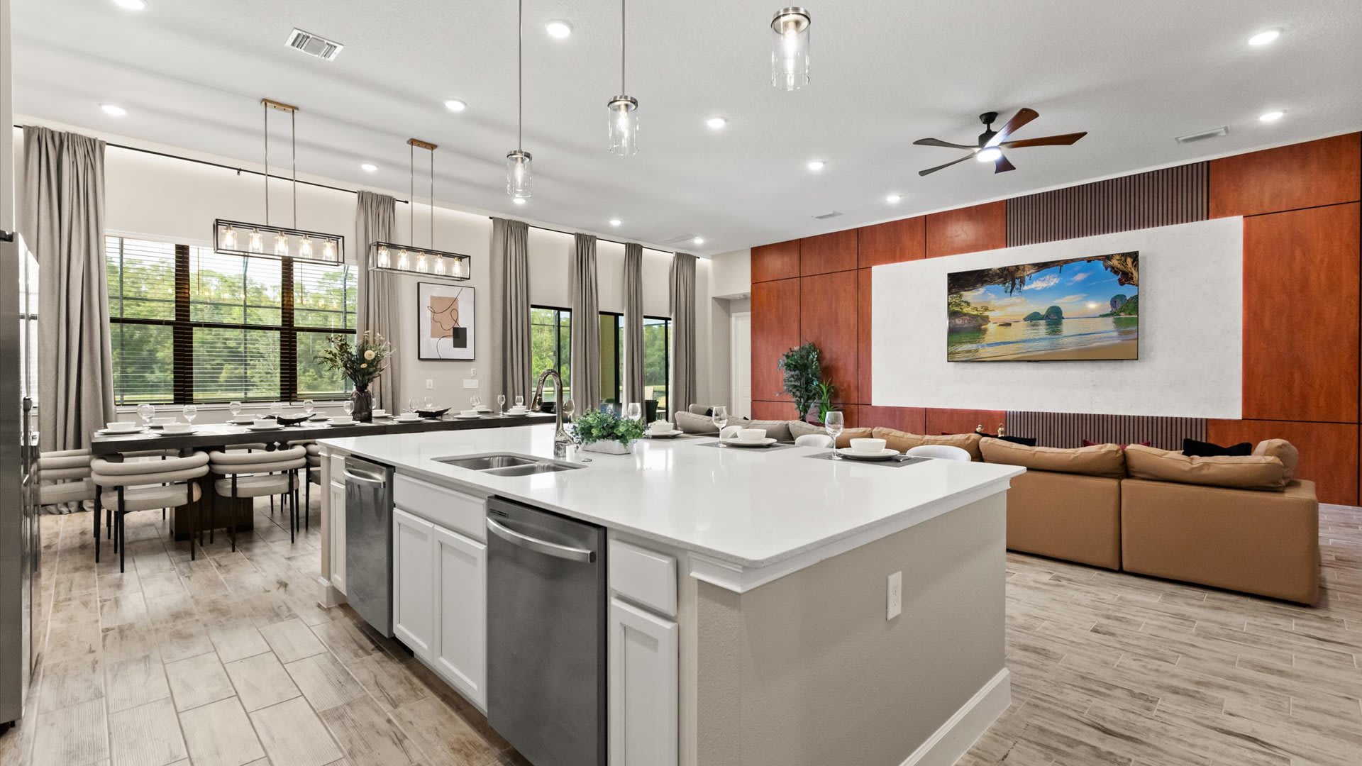 Expansive open-concept space showcasing a modern kitchen with a large island, flowing into a dining area with a long table, and a living room with a sectional sofa. Large windows overlook the exterior.