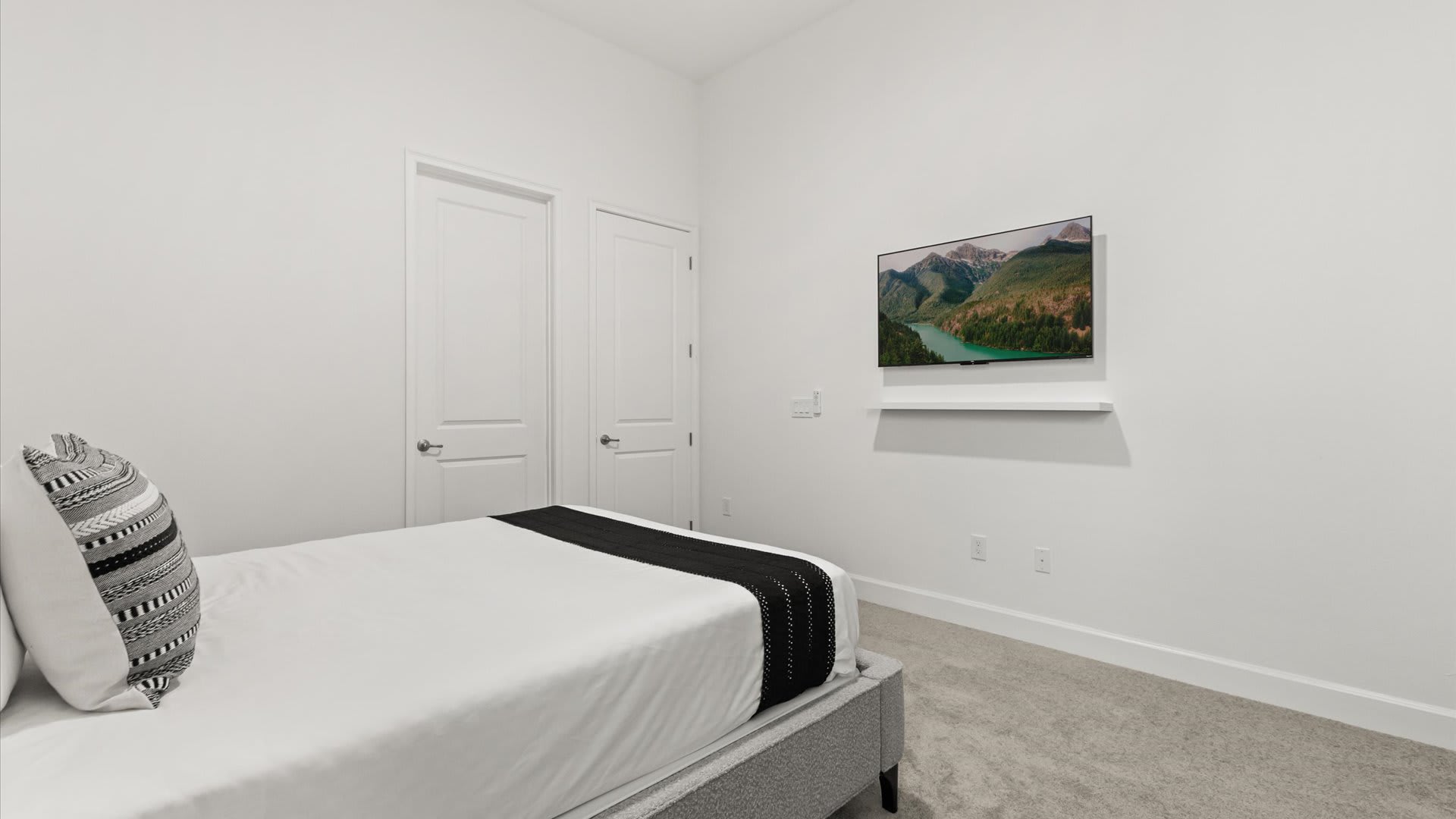 A minimalist bedroom with white walls, a bed featuring white and black bedding, and a wall-mounted TV displaying a mountain lake scene. Two white doors are visible on the left, and the floor is carpeted.