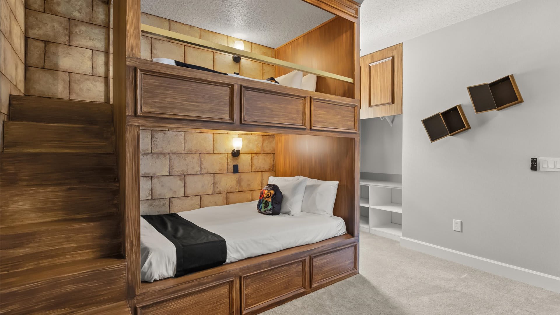 Themed bunk bed room with dark wood bunk beds, stone-look wall panels, and a small staircase leading to the top bunk. The lower bunk has a white mattress with black and colorful pillows.