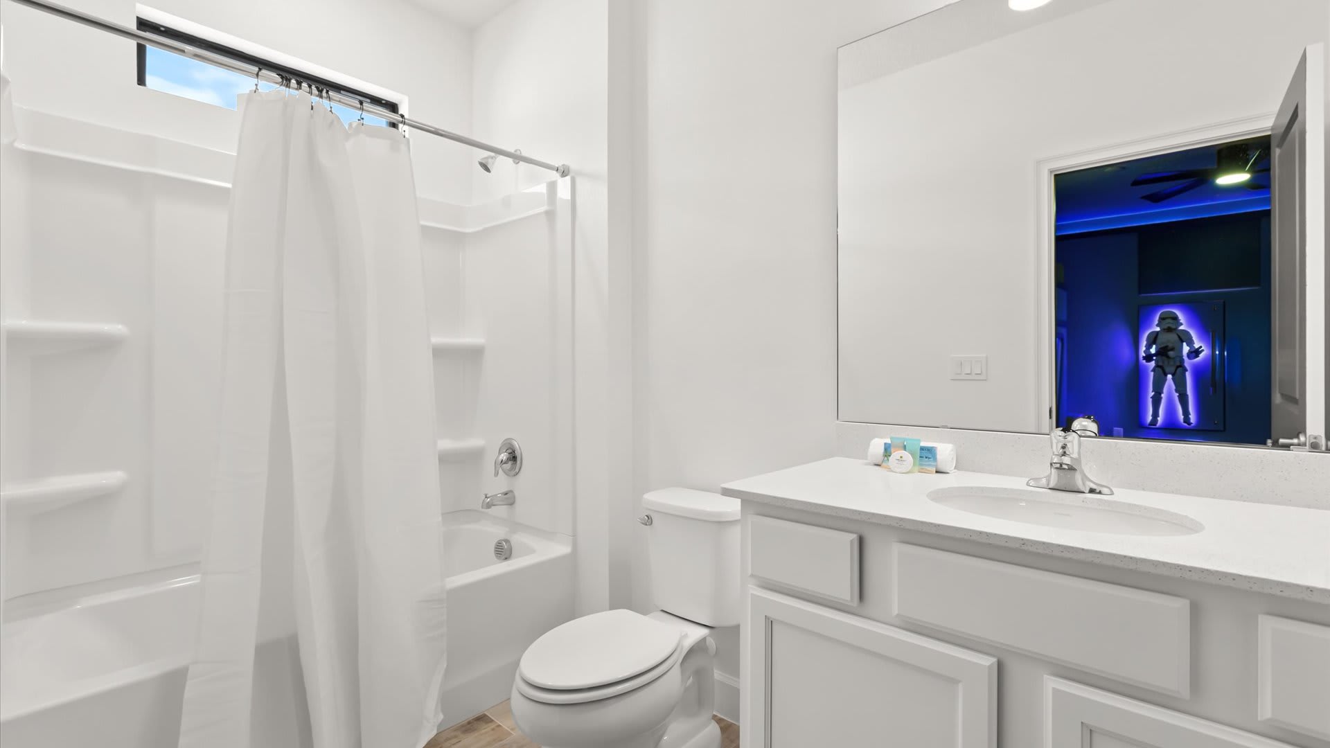 Clean white bathroom with a single vanity, white countertop, and a shower/tub combo with a white curtain. The mirror reflects the blue-lit Star Wars themed bedroom with its Stormtrooper decal.