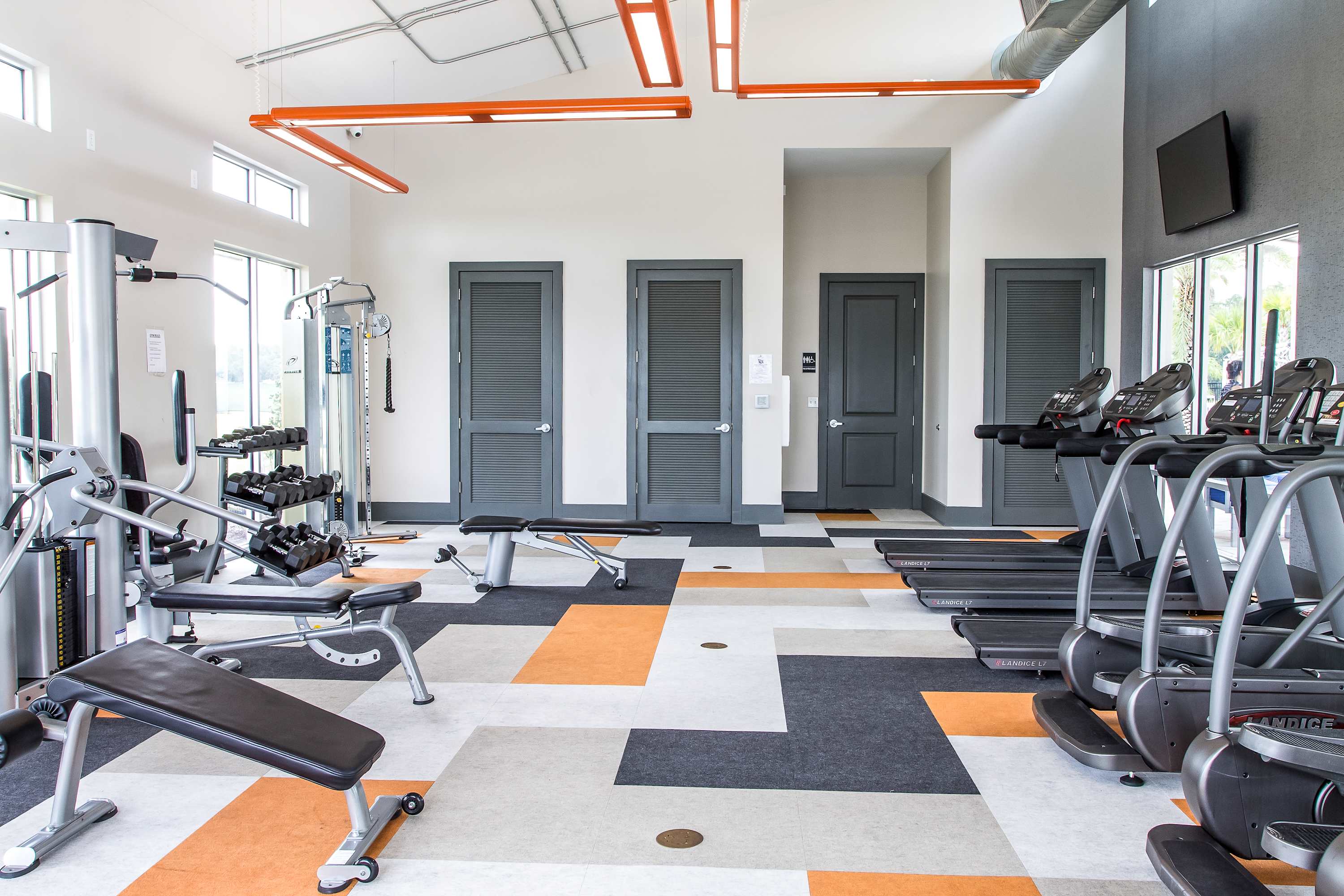 Spacious fitness center with various exercise machines including treadmills, weight benches, and a multi-station gym. The floor features a modern geometric pattern in orange, gray, and white.