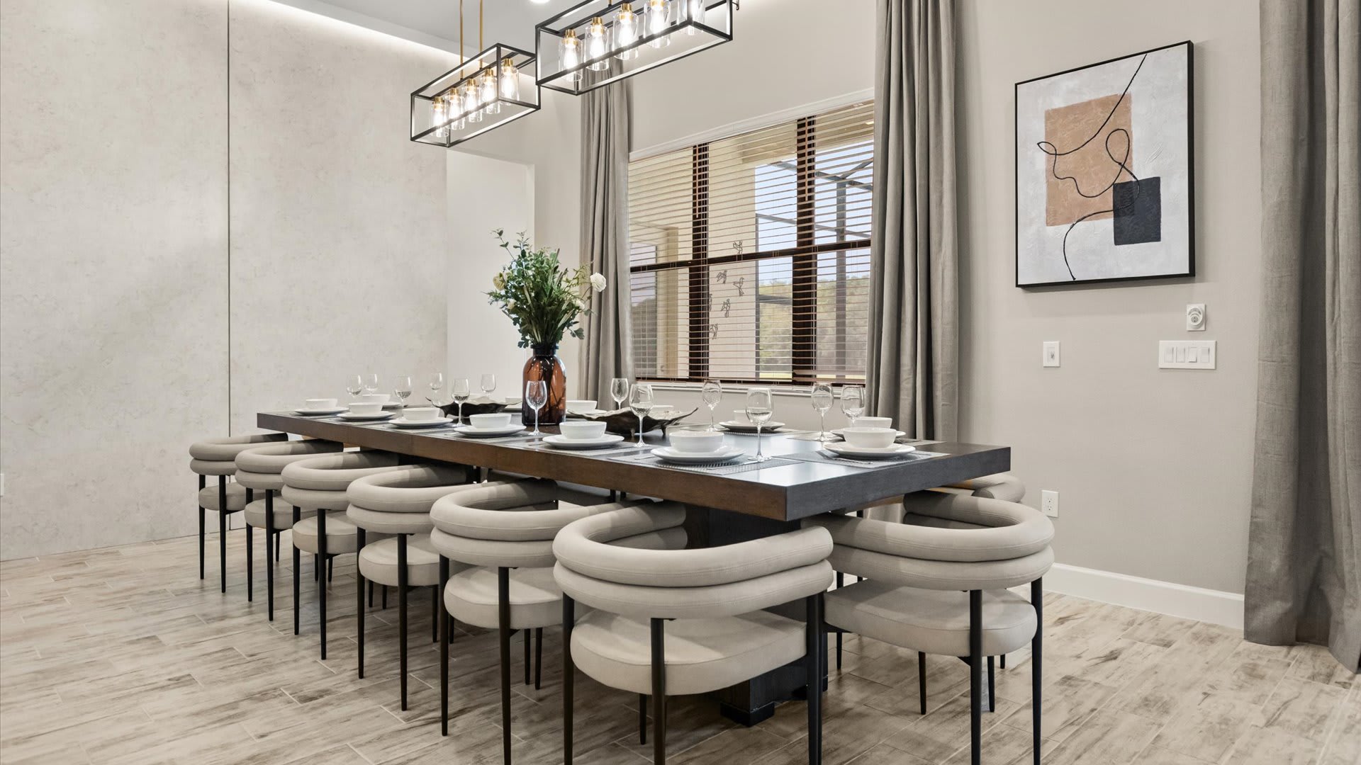 A long, modern dining table with a dark finish, surrounded by light-colored, curved upholstered chairs, set for a large group. Two linear chandeliers illuminate the space, with large windows providing natural light.