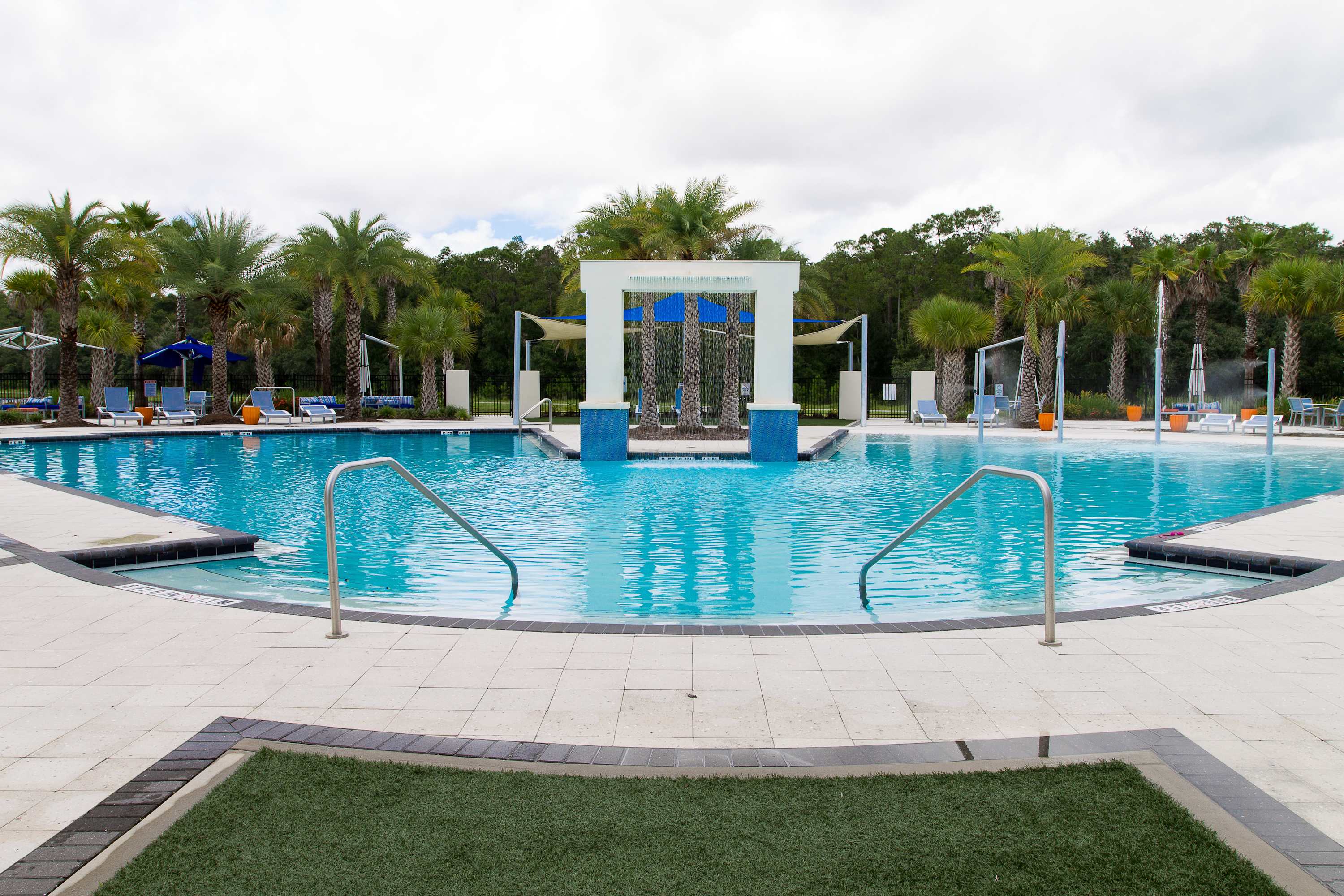 Expansive communal swimming pool with a shallow entry area and water features, surrounded by palm trees and lounge seating. A large white archway stands at the pool's edge.