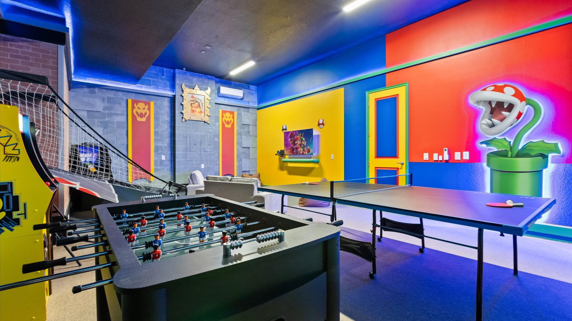 A colorful game room features a vibrant Super Mario Bros. themed mural with Bowser banners and a large Piranha Plant. The room includes a foosball table, a ping pong table, and an arcade basketball game, all under blue accent lighting.