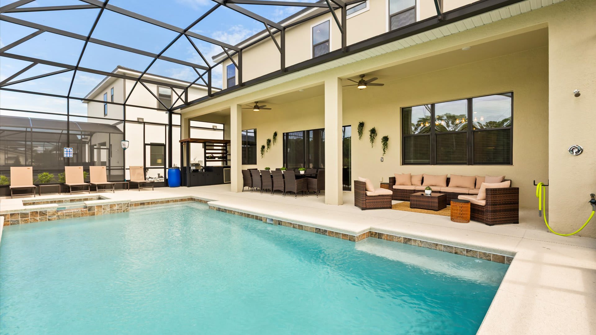 A spacious screened-in outdoor area features a large swimming pool and a covered patio with a dining table and a comfortable seating area. The patio is furnished with dark wicker furniture and overlooks the pool and surrounding landscape.