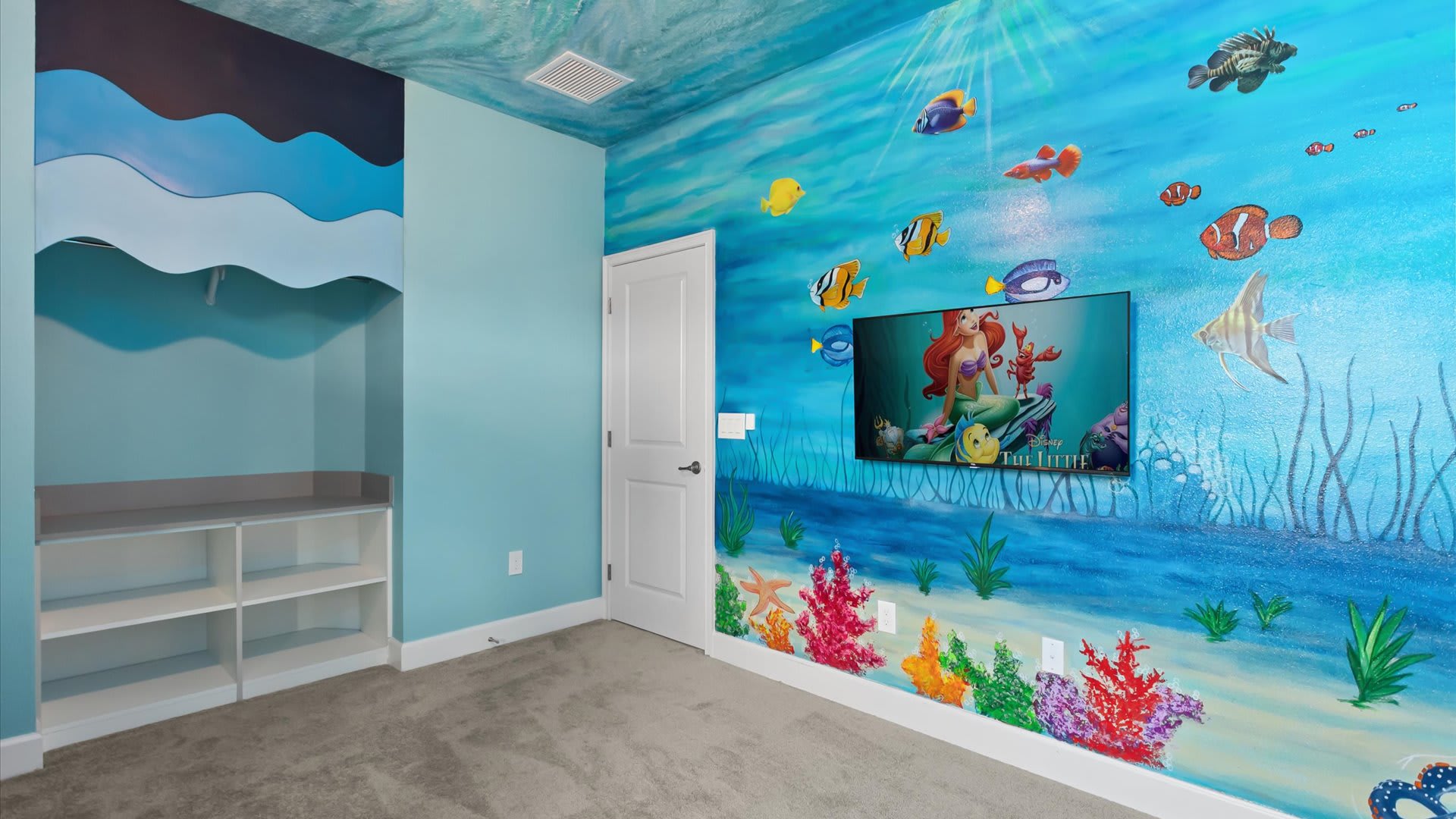 A vibrant 'Little Mermaid' themed bedroom features a large mural of Ariel and sea creatures on a blue wall, a wall-mounted TV, and a built-in storage unit with shelves. The ceiling is also painted with an underwater theme.