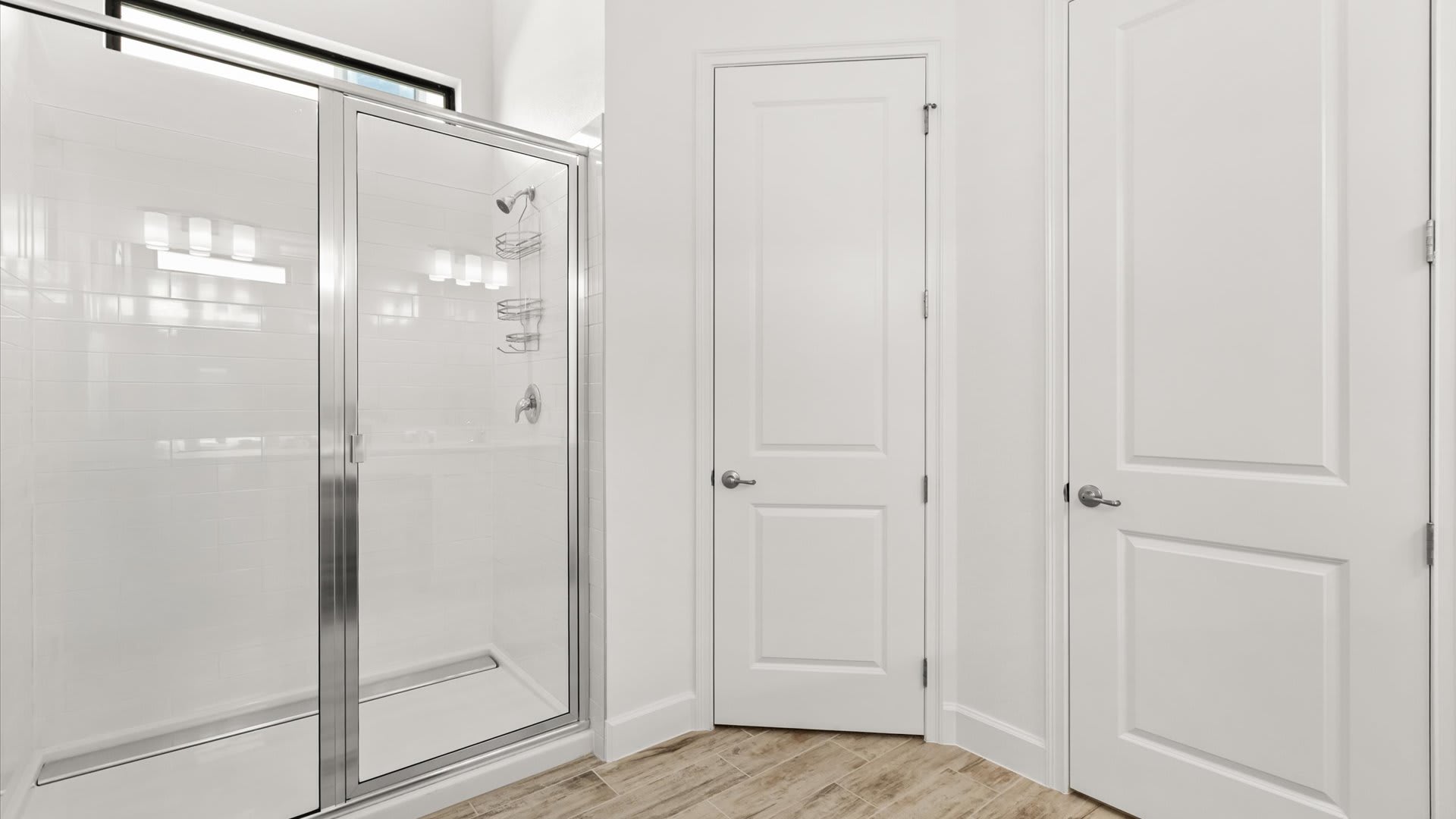 Modern glass-enclosed shower with white subway tile walls, chrome fixtures, and a built-in shelf. Two white panel doors are visible next to the shower, set against a wood-look tile floor.
