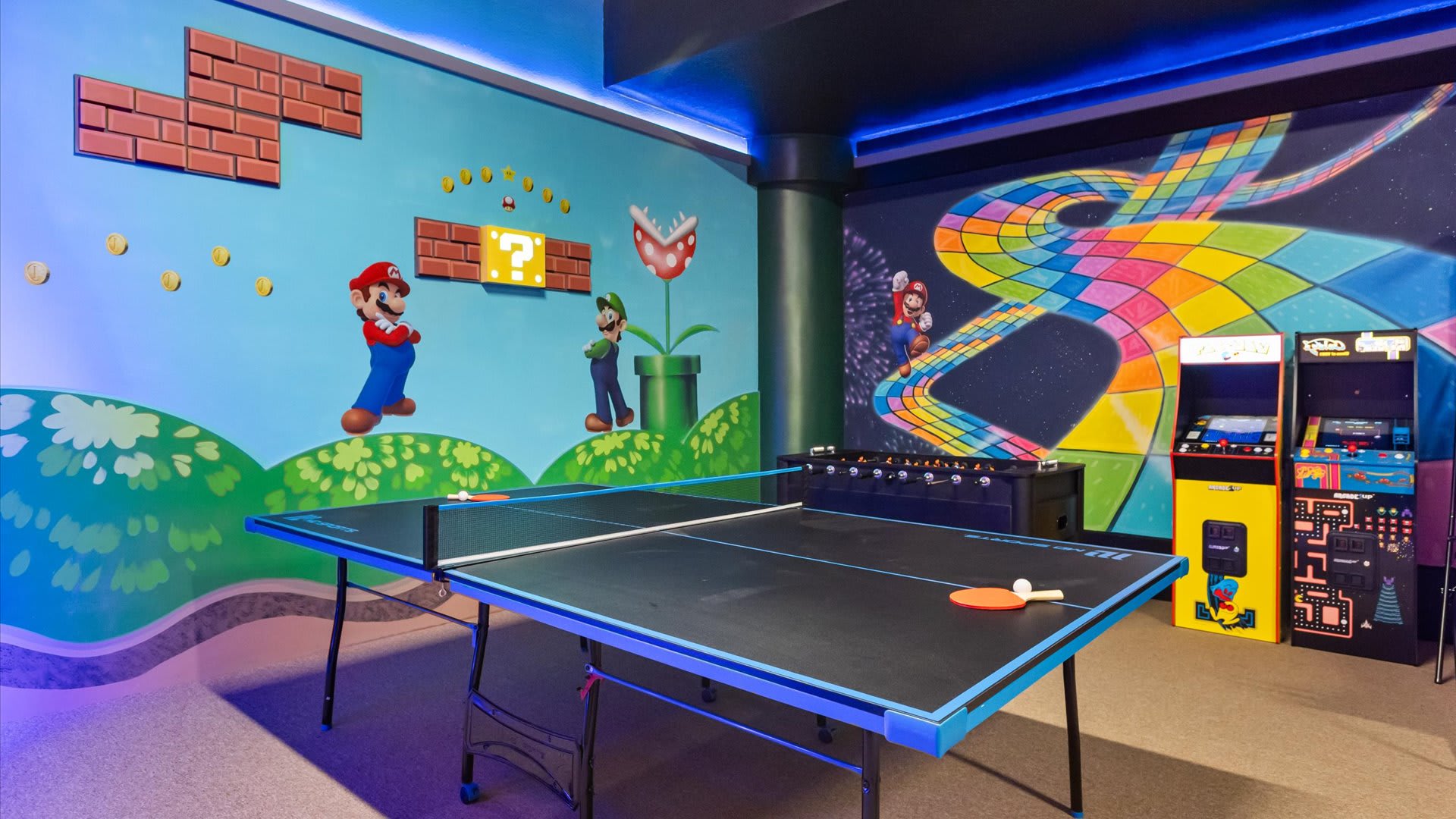 A vibrant game room features a Super Mario Bros. themed mural on the walls, including Mario, Luigi, and Piranha Plants. A black ping pong table with blue accents is in the foreground, with arcade machines and a foosball table in the background.