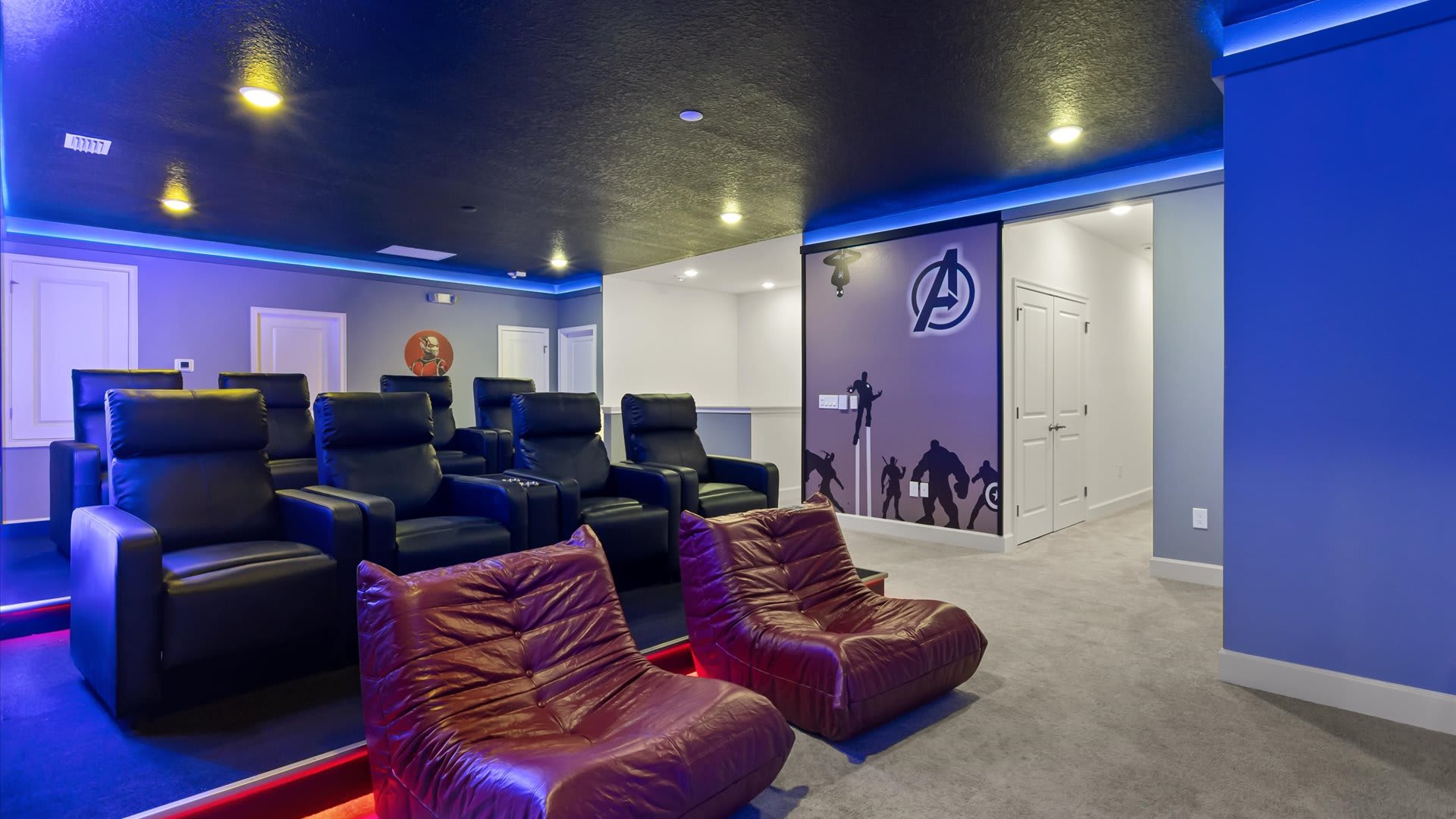 A home cinema room with multiple rows of black leather recliners, two red beanbag chairs, and blue accent lighting along the ceiling and floor. A wall mural with the Avengers logo and characters is visible on the right.