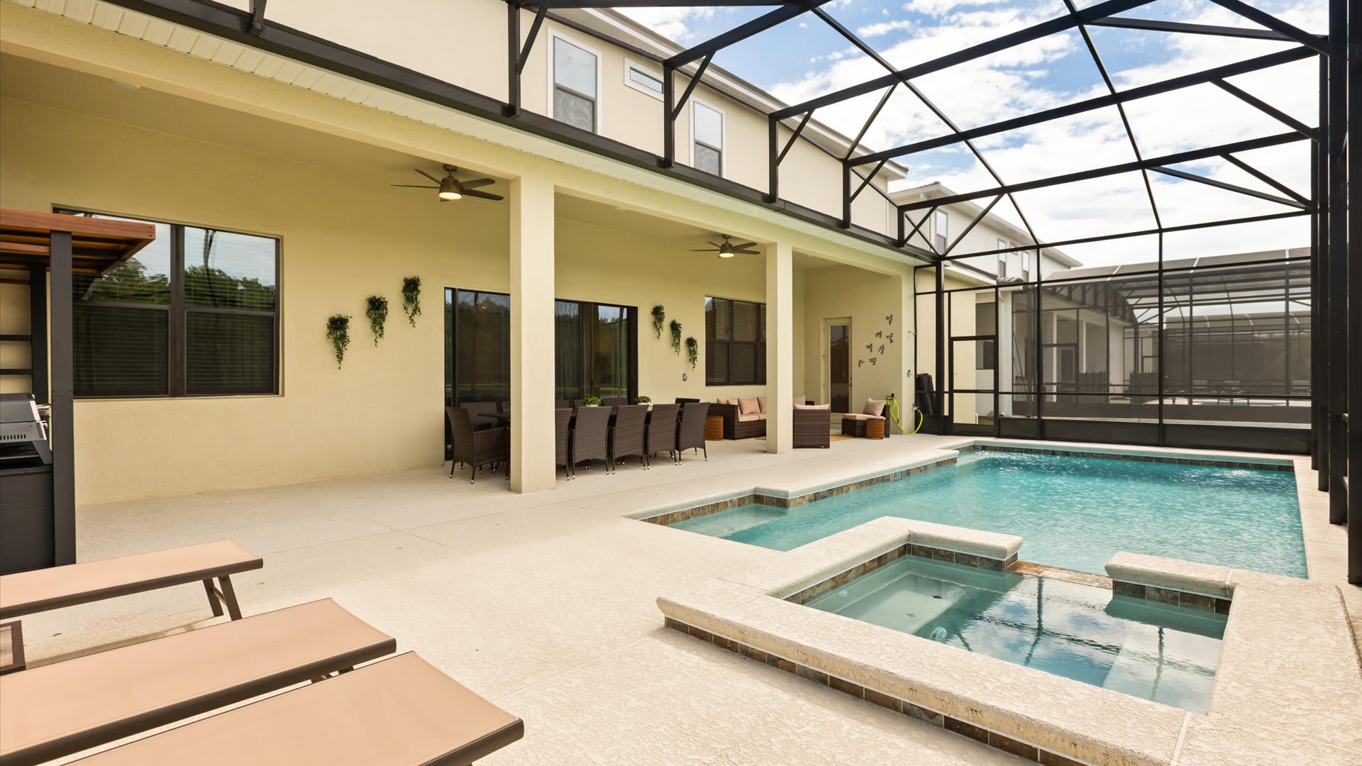 A wide view of the screened-in pool area shows a large swimming pool with an integrated hot tub, surrounded by a light-colored deck. A covered patio area with outdoor seating and lounge chairs is visible, with the house exterior in the background.