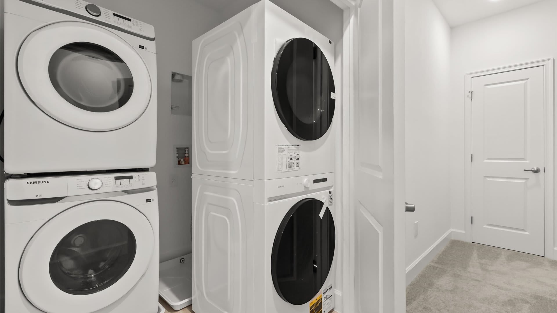 Modern laundry area with two sets of white stacked Samsung washer and dryer units. The room has light gray walls and a light-colored carpet, with a closed white door in the background.