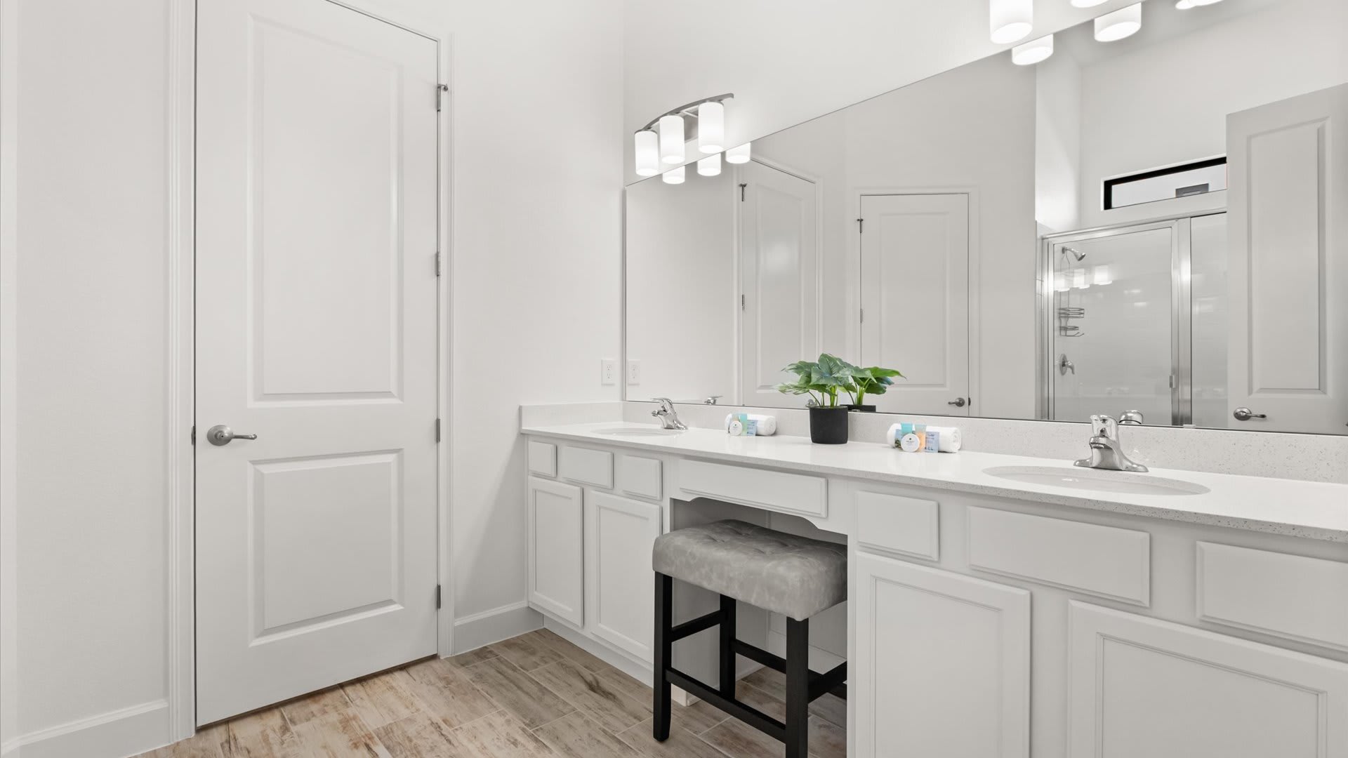 Bright white bathroom featuring a double vanity with white quartz countertops, chrome fixtures, a large mirror, and a dedicated makeup area with a stool. A white panel door is visible on the left.