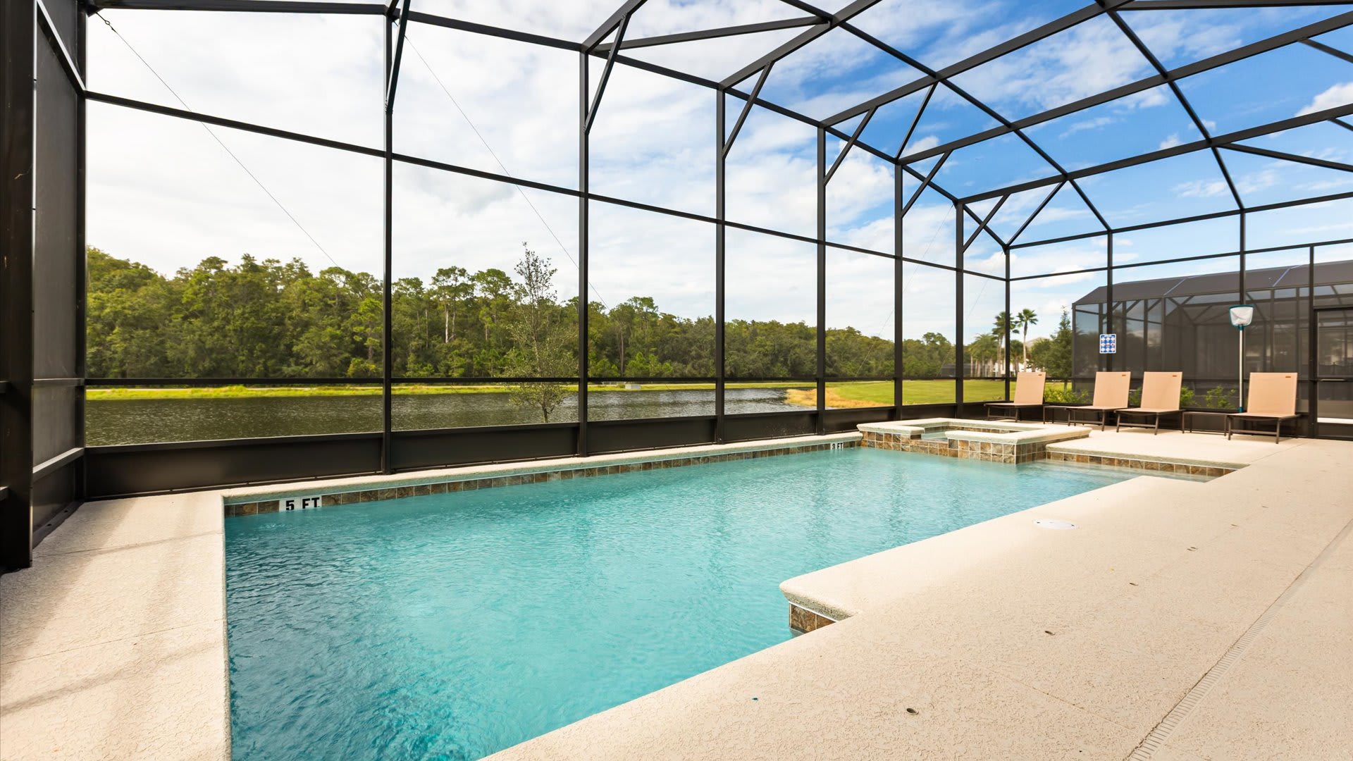 A large screened-in private pool with an integrated hot tub is set against a lush green landscape and a lake under a partly cloudy sky. The light-colored pool deck includes a shallow entry area and distant lounge chairs.