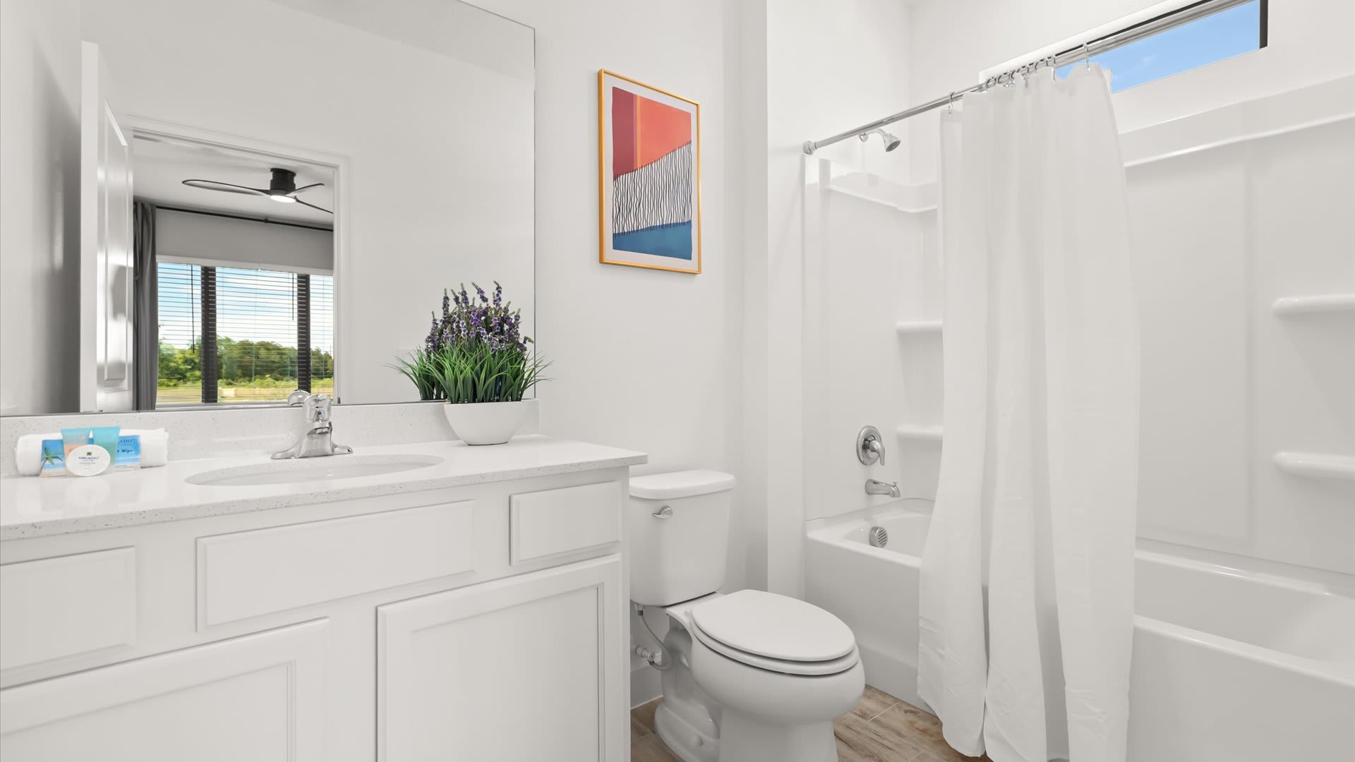 Bright white bathroom featuring a single vanity with a white quartz countertop, a chrome faucet, and a shower/tub combo with a plain white curtain. A mirror reflects a room with a ceiling fan and horizontal blinds.
