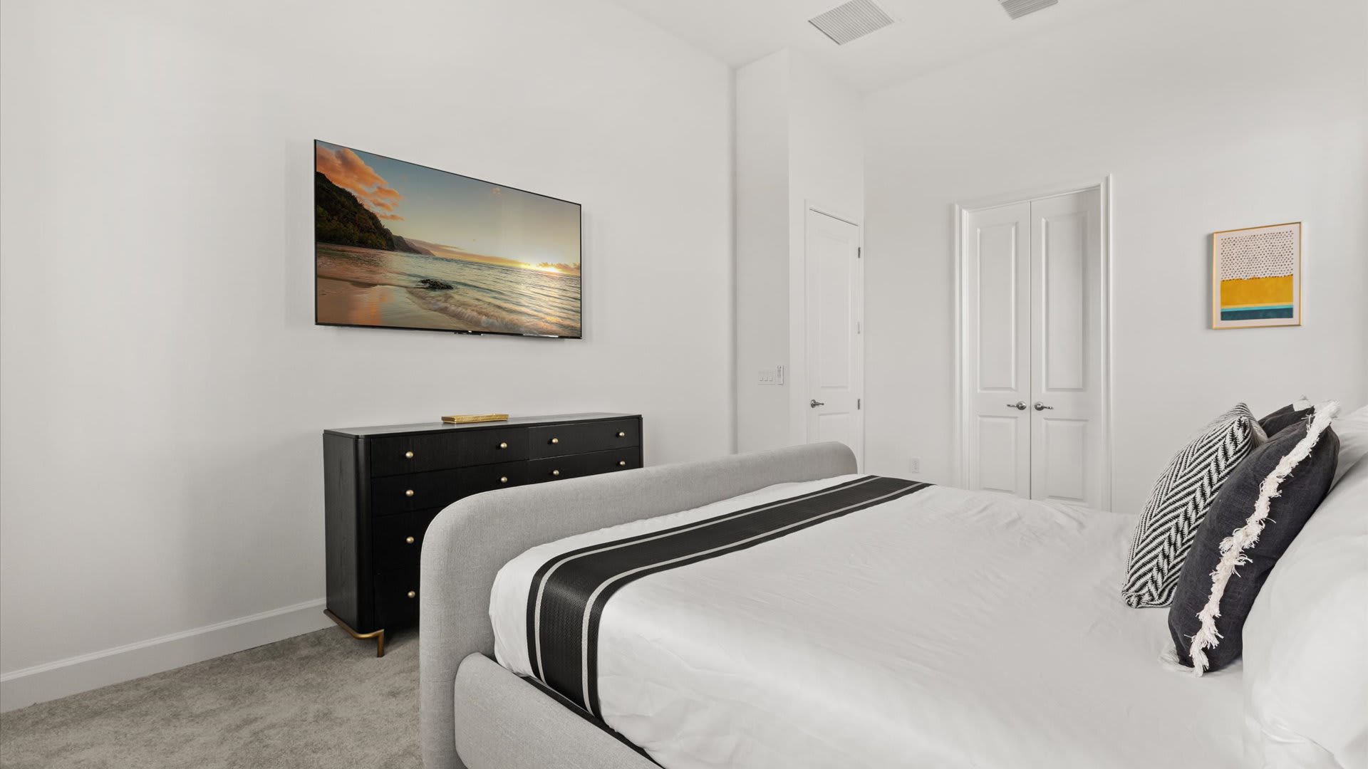Contemporary bedroom with a light gray upholstered bed and black and white bedding, facing a wall-mounted TV above a black dresser. The room has light gray carpet and white walls.