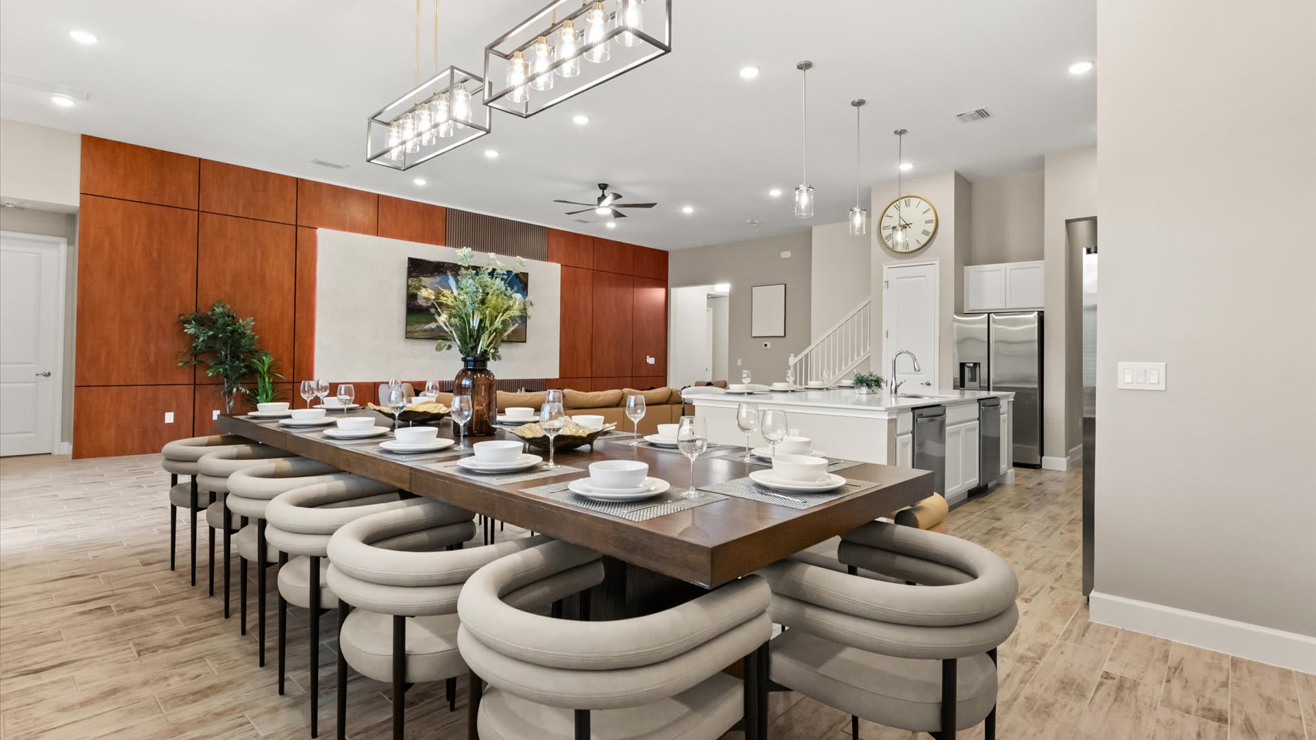 A long, modern wooden dining table with twelve upholstered chairs is set for a meal, illuminated by two contemporary rectangular chandeliers. The dining area is open to a kitchen with a large island and a living area with a sectional sofa in the background.