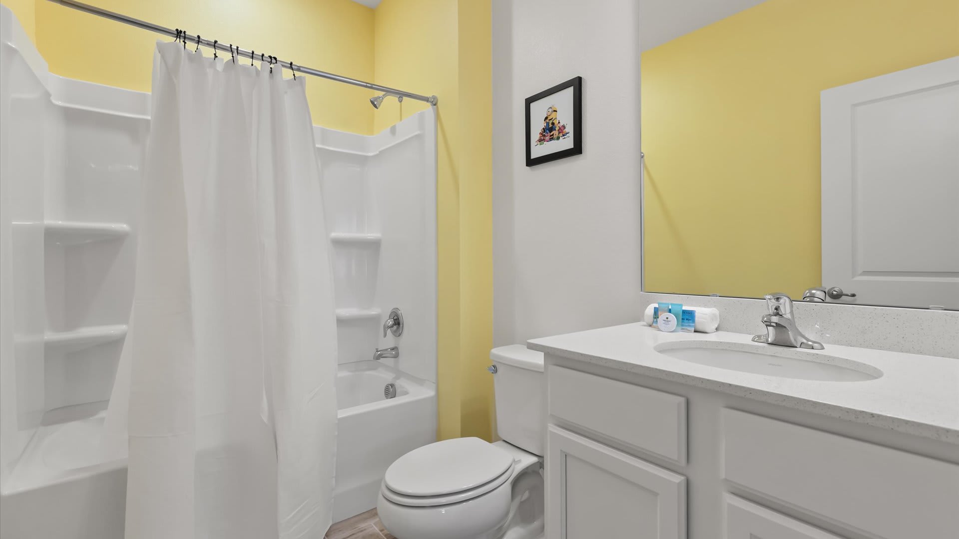 A bright bathroom features a yellow accent wall, a white vanity with a single sink, and a large mirror. It includes a white toilet and a shower/tub combo with a white curtain.